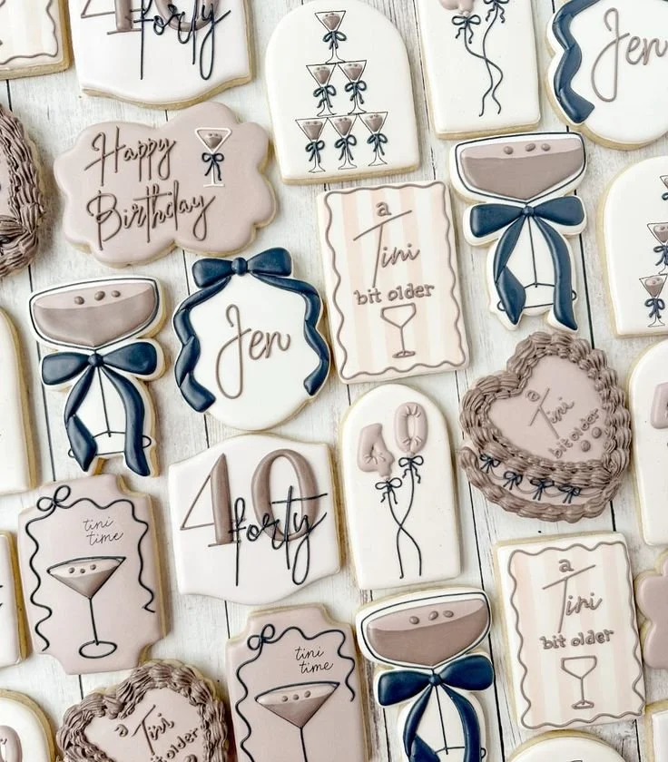 Decorative cookies with birthday and celebration themes, featuring phrases like 'Happy Birthday', '4 holy', and 'a tiny bit older'. Some cookies have balloon and champagne glass designs, decorated with bows and scalloped edges.