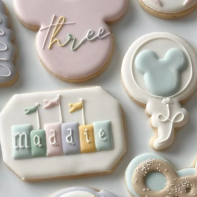 Decorated cookies with pastel icing for a birthday celebration, including a cookie with the name 'Maddie,' a pink heart-shaped cookie with the word 'three,' and other assorted cookies.