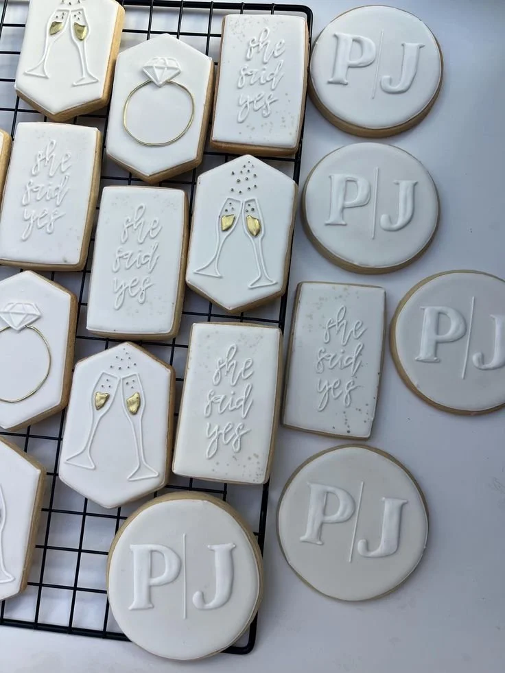 Wedding-themed cookies on a cooling rack with white icing and gold details, featuring designs of champagne glasses, a diamond ring, and the message 'she said yes,' along with cookies with the initials 'PJ'.