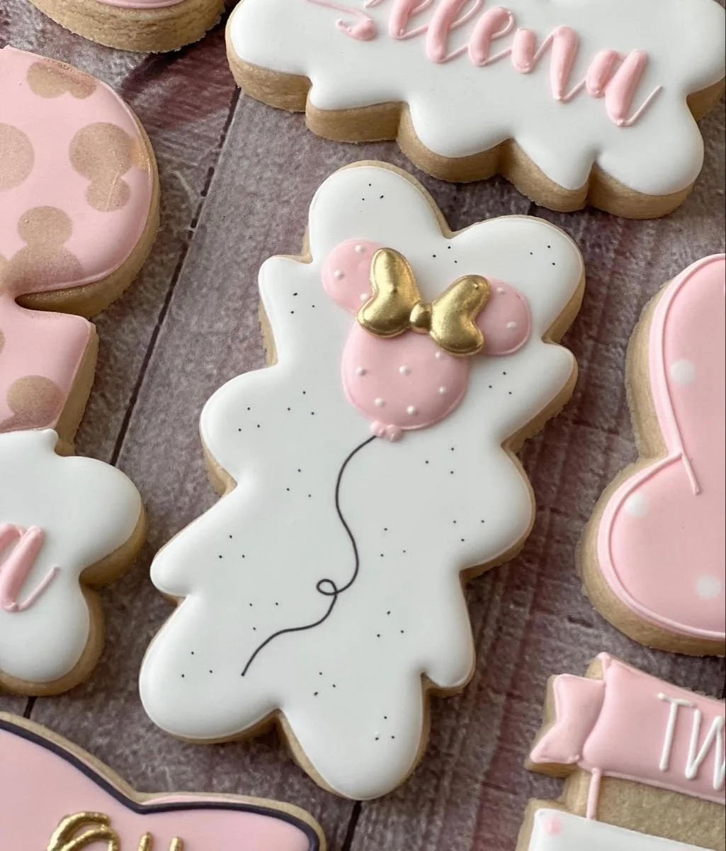 Decorated cookies on a wooden surface, including a white cookie shaped like a bunny with pink and gold butterfly decoration and black speckles, and other pink and white cookies with various designs.