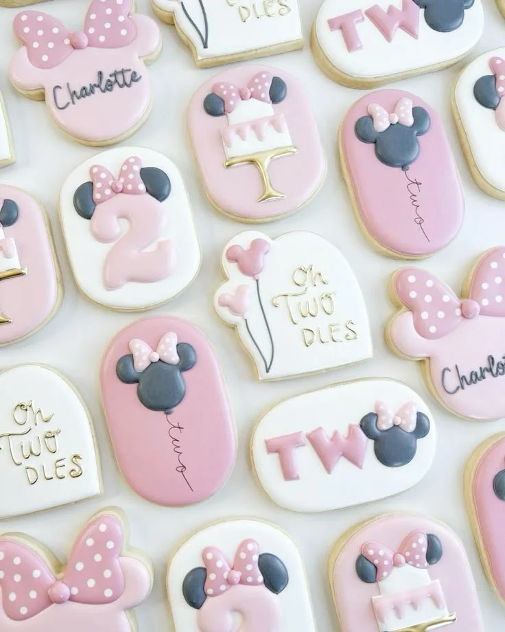 Decorated sugar cookies for a Disney-themed celebration, featuring Minnie Mouse ears, pink bows, text like 'Oh Two DIES', numbers '2', and Minnie Mouse silhouettes, colored in pink, white, black, and gold.