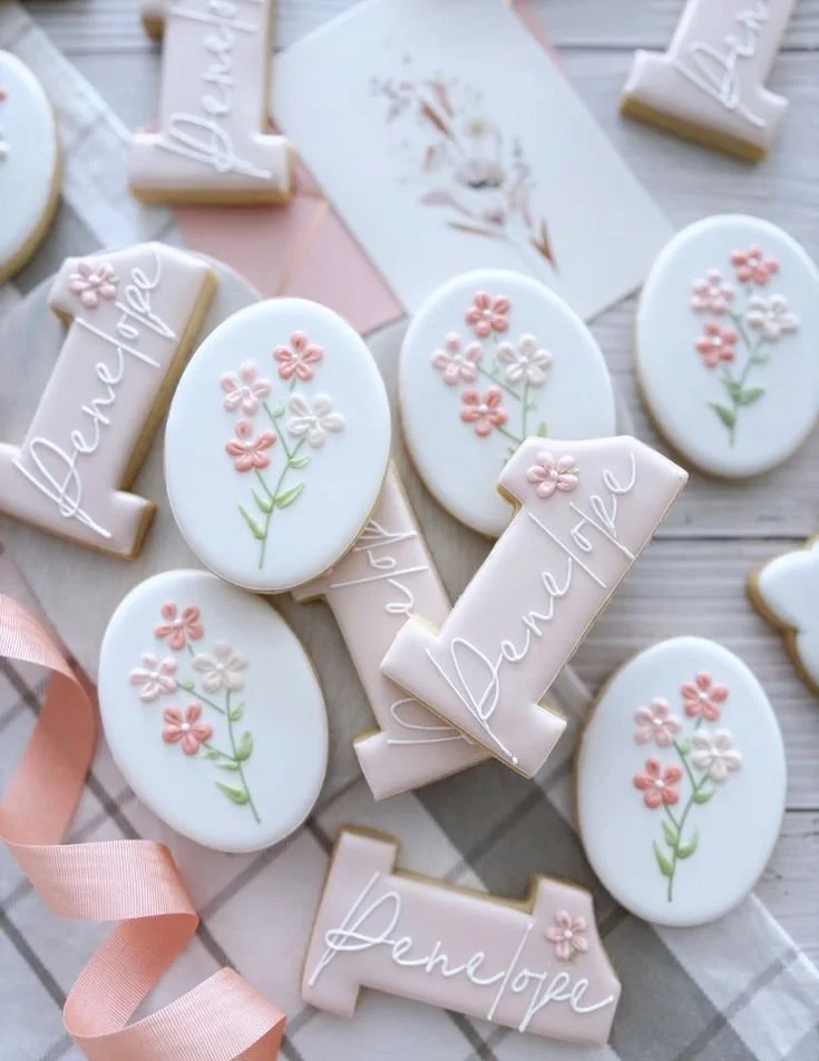 Decorated cookies with pastel icing featuring flowers, the word "Dance" on one cookie, and the word "Penteope" on others, arranged on a light wooden surface with pink ribbon.