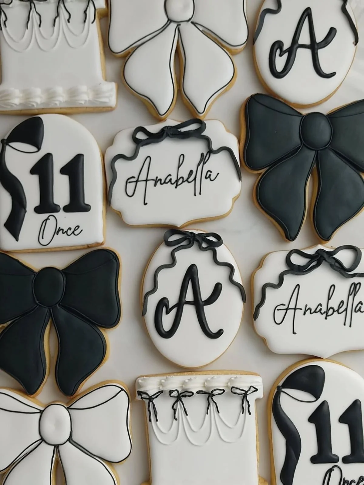 Decorative cookies with black and white themes, featuring bows, initials, numbers, and the name Anabella, likely for a celebration.