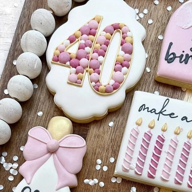 Decorated cookies and cake for a 40th birthday, including a large cookie with '40' filled with pink, purple, and beige candies, a pink bow-shaped cookie, and other birthday-themed cookies with candles and messages, all arranged on a wooden surface wi