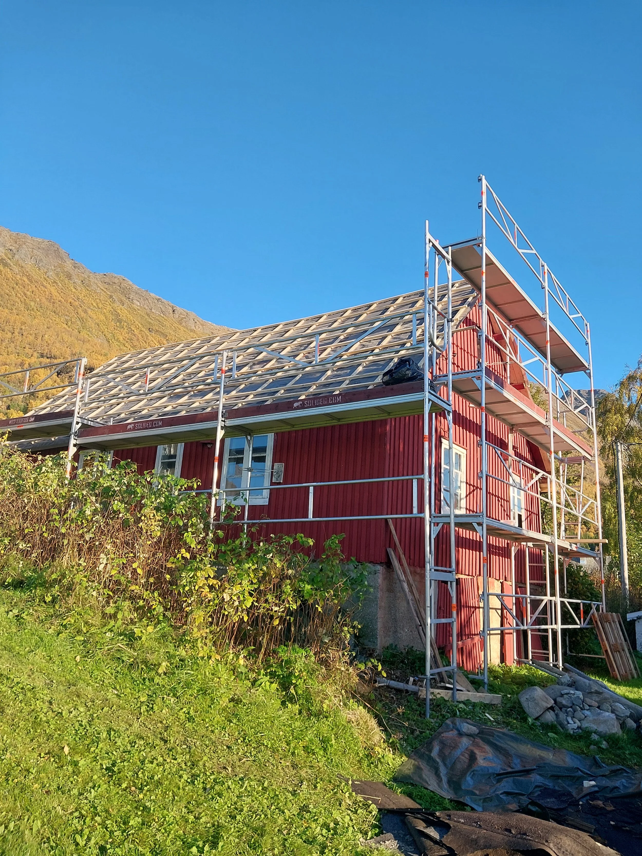 En rød fjøs under bygging med stillas rundt, taket er under oppføring, og det er en bakgrunn av fjell og blå himmel.