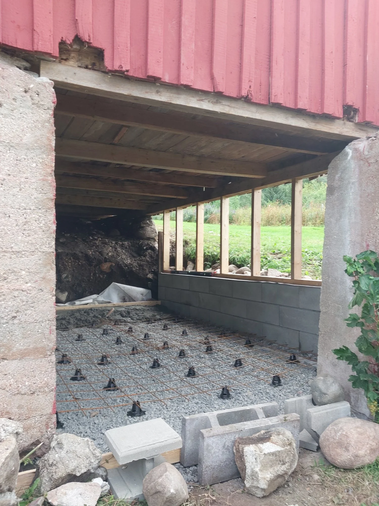 Påbegynt utendørs konstruksjon under en bygning, med grus, armeringsjern og murblokker, hvor fundamentet oppgraderes.