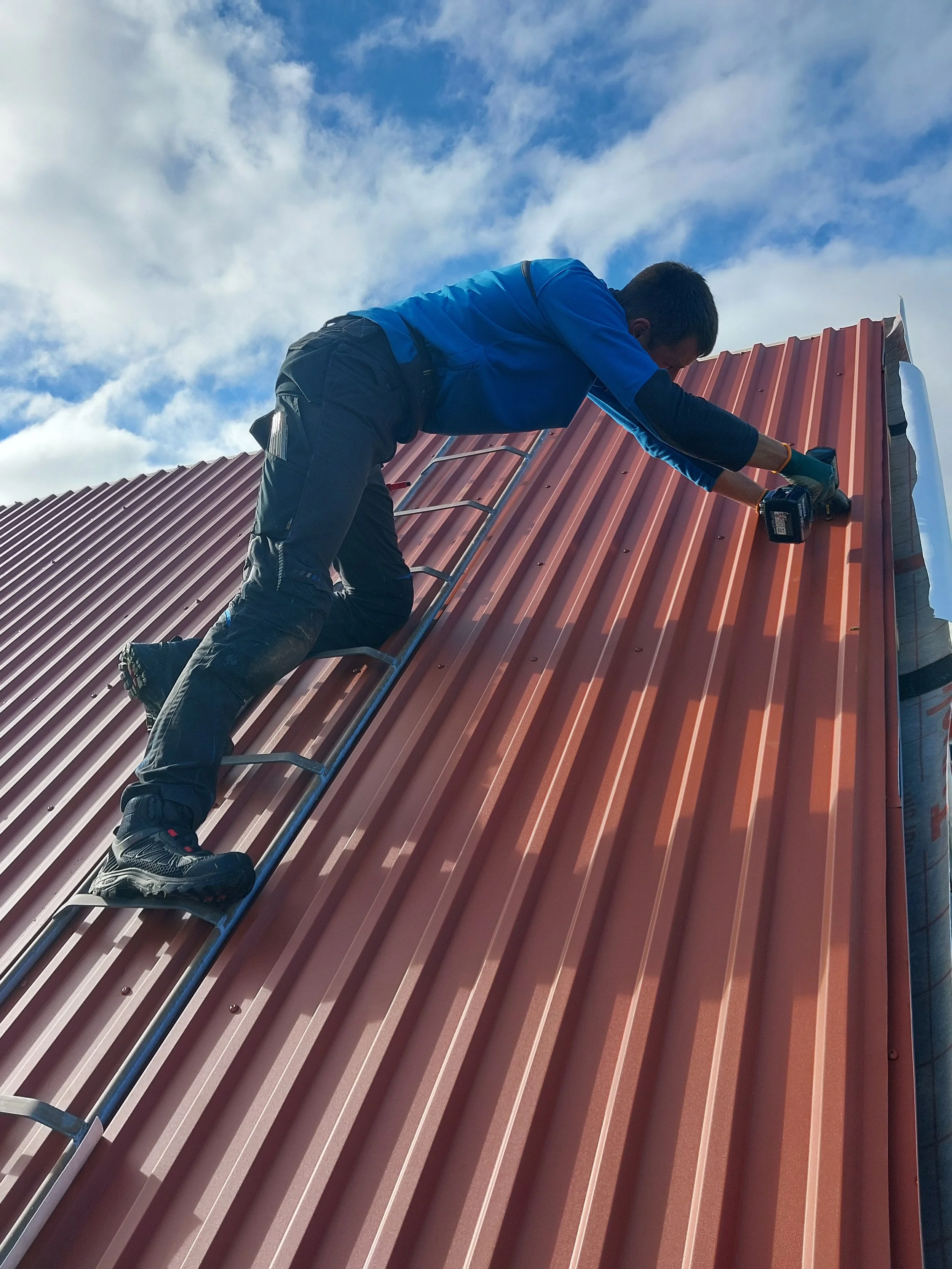 En person som jobber på et tak med røde metallplater, bruker verktøy og er iført jobbklær under en klar blå himmel med noen skyer.