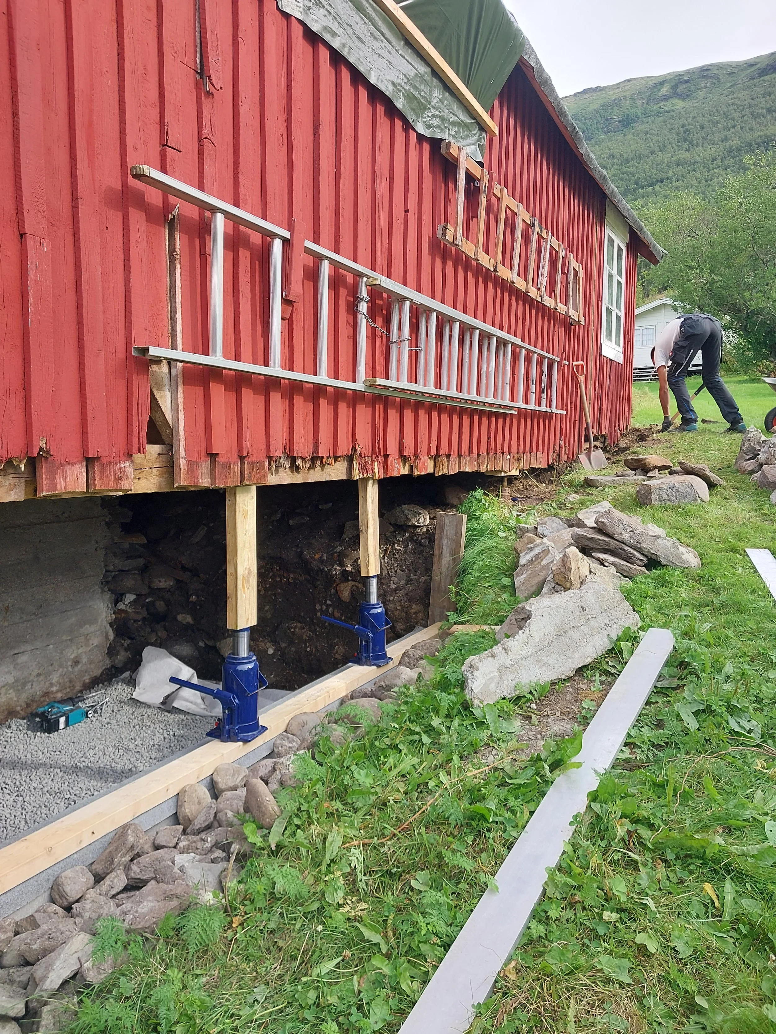 En byggeplass foran en rød fjøs, der det pågår restaureringsarbeid. To personer er i ferd med jekking.