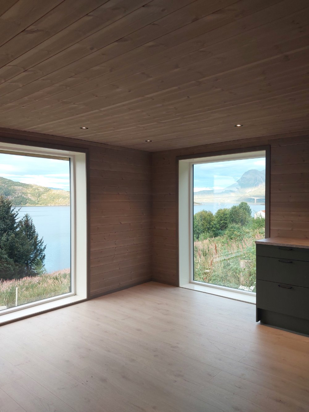 Et tomt rom med store vinduer som viser utsikt over sjø og fjell, innvendige trevegger og tak, og en gulv av lyst tre.