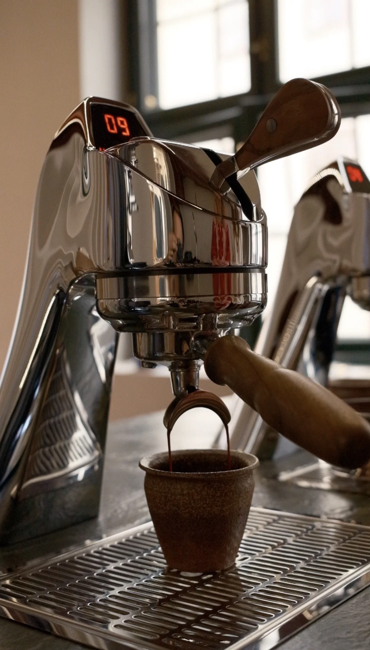 En espressomaskine, der laver espresso i en kop. Der er en digital skærm på maskinen, der viser tallet 09. Maskinen er i rustfrit stål, og baggrunden viser et vindue.