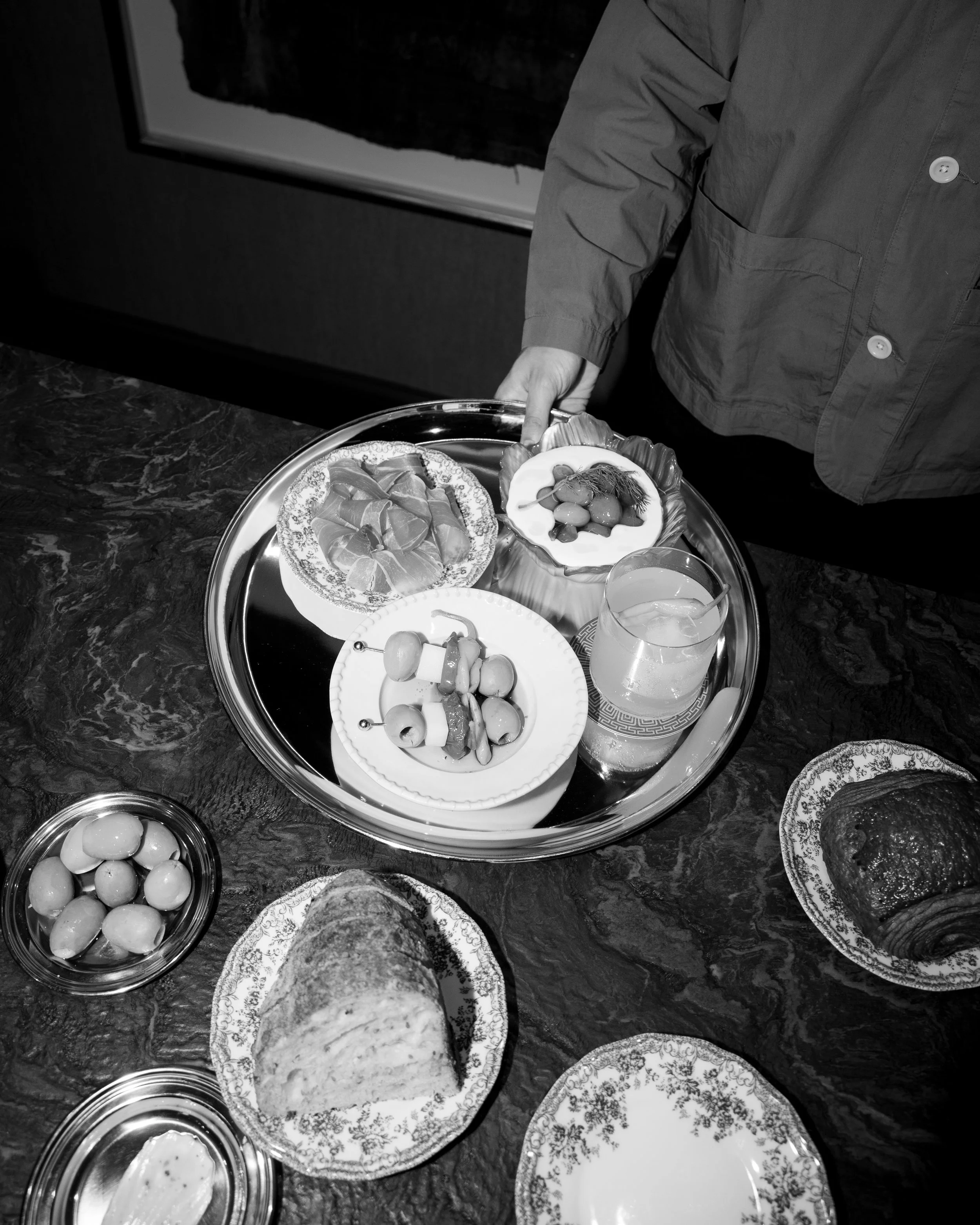 Billede af et serveringsbord med forskellige anretninger, herunder skinke, brød, oliven, snacks og drikkevarer, på en bordflade af natursten.