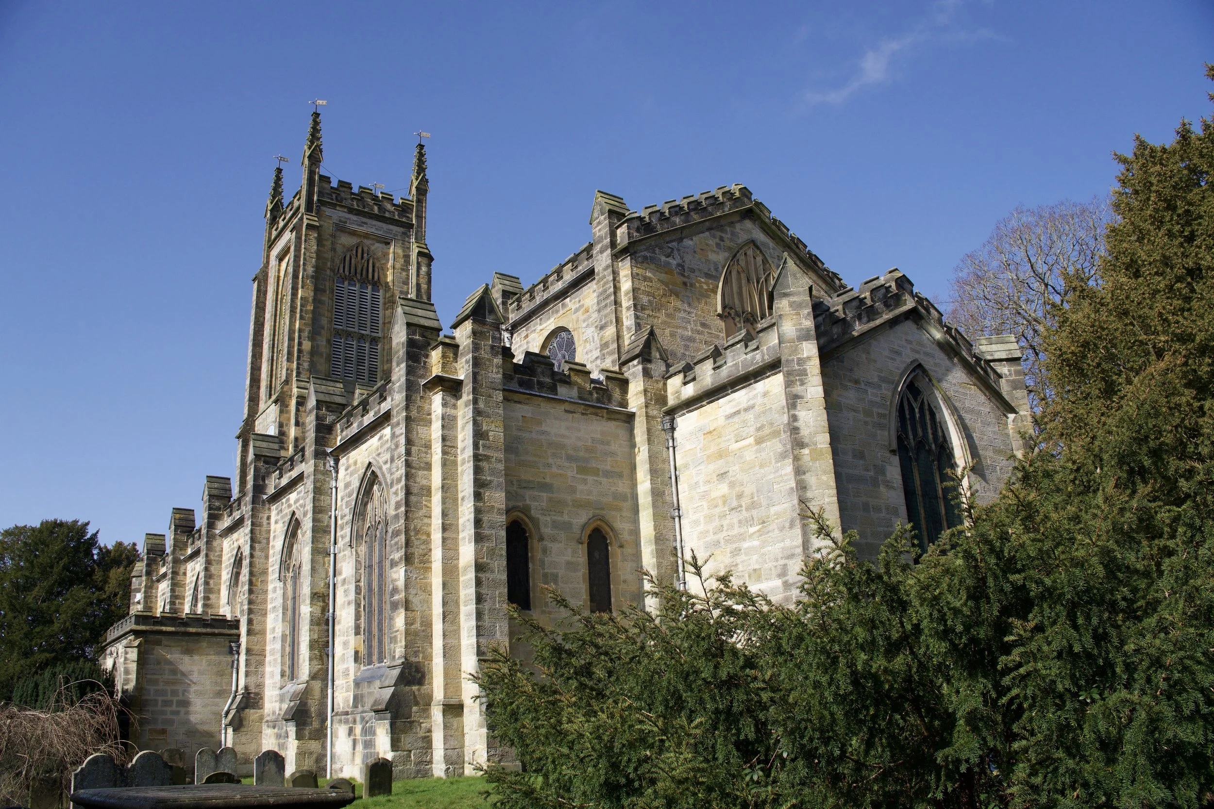 A stone Gothic-style church with pointed arched windows and a tall bell tower, surrounded by trees and bushes, under a clear blue sky which is the St Swithun East Grinstead church building.
