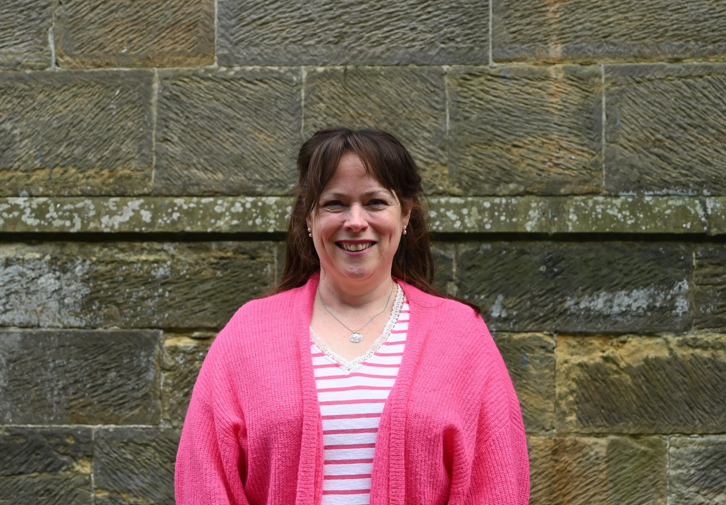 A woman wearing a pink sweater smiles in front of a stone wall.