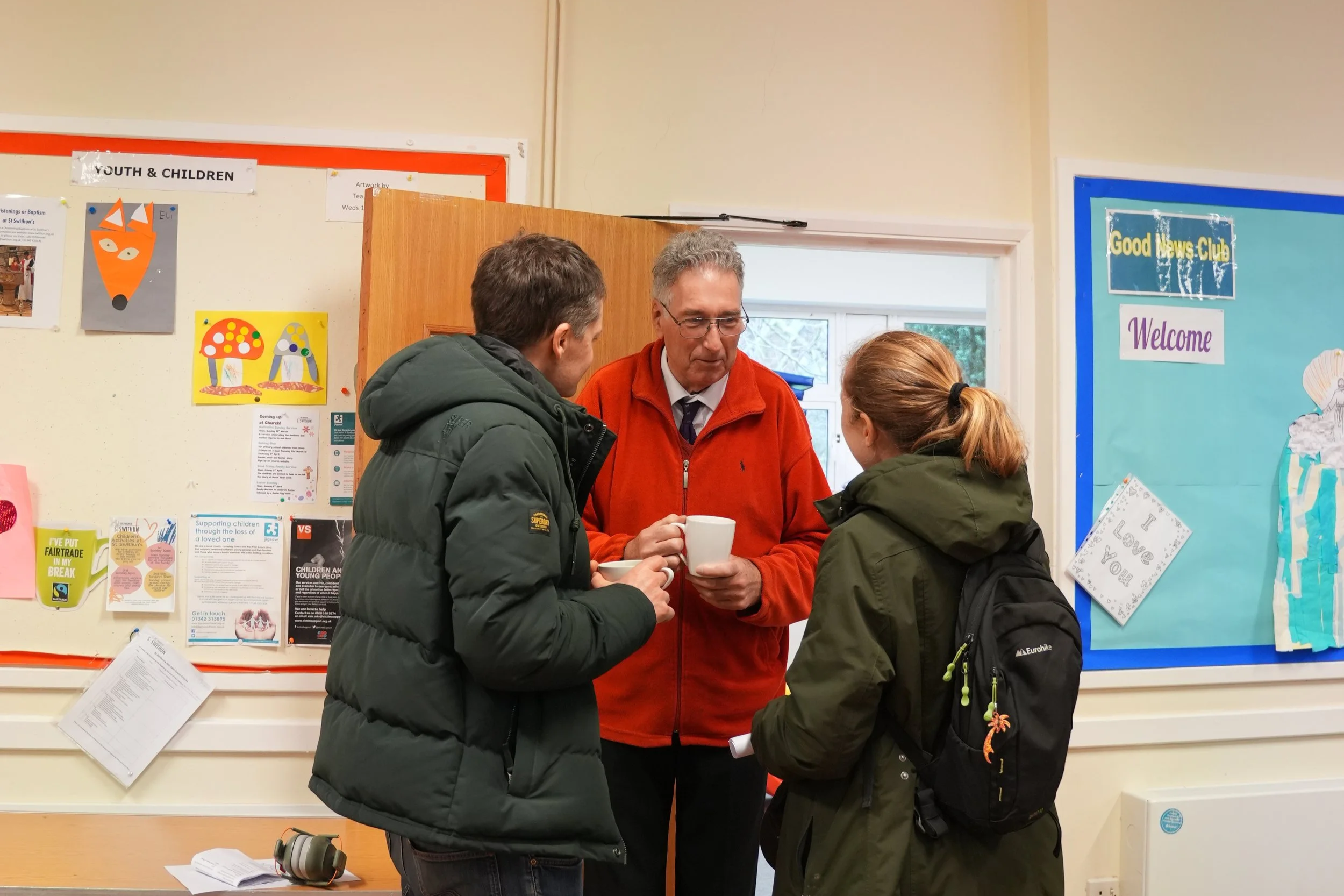 Three people engaged in conversation inside a community center, two are wearing warm jackets, and one man in the middle, wearing glasses and a red jacket, holding a white mug.