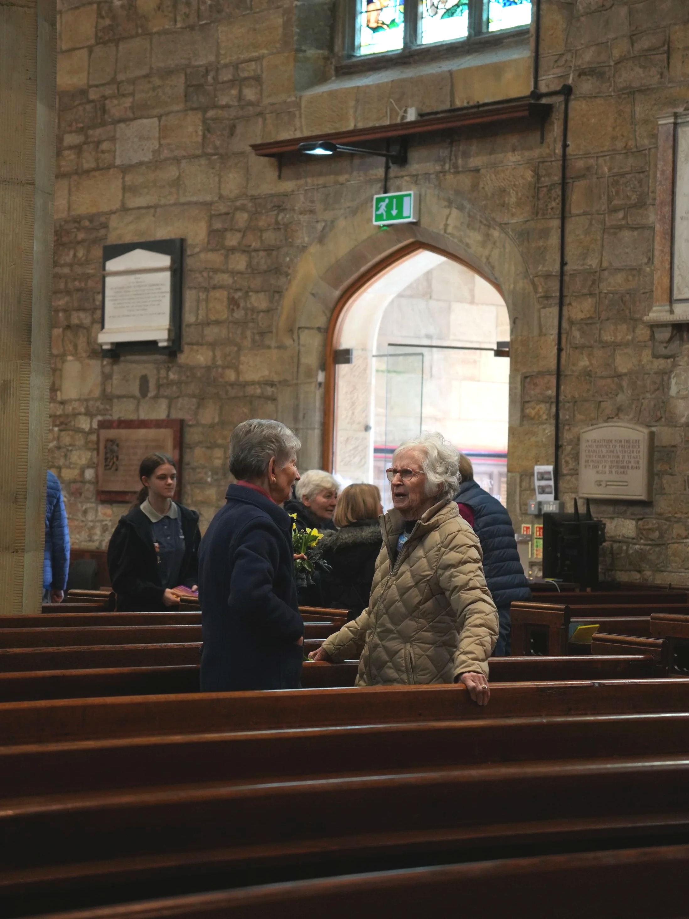 Inside a church with exposed stone walls, several elderly women are engaged in conversations, one holding flowers. Wooden pews are visible, and a stained glass window is at the top. There is an emergency exit sign above an arched doorway.