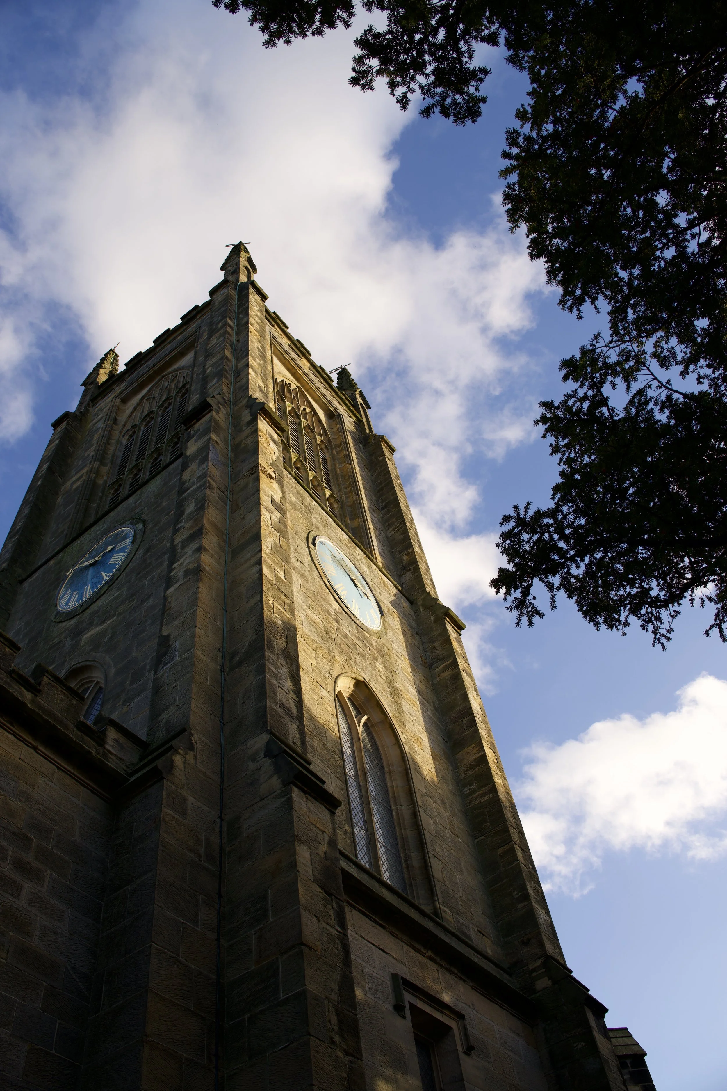 Low-angle view of a stone church clock tower against a partly cloudy sky with trees in the foreground.
