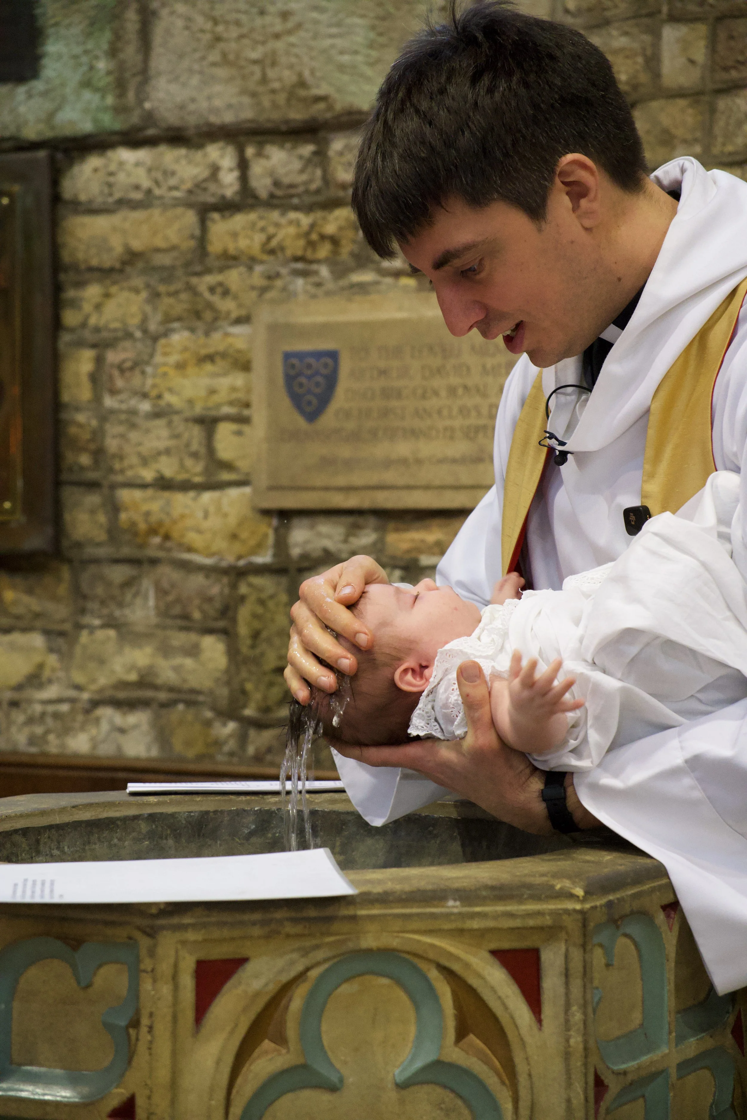 A young man in white robes with a gold sash baptizing a baby in a church setting. The man has water on his head and is smiling down at the baby, who is dressed in a white outfit and lying in a baptistery with a pipe in the background.