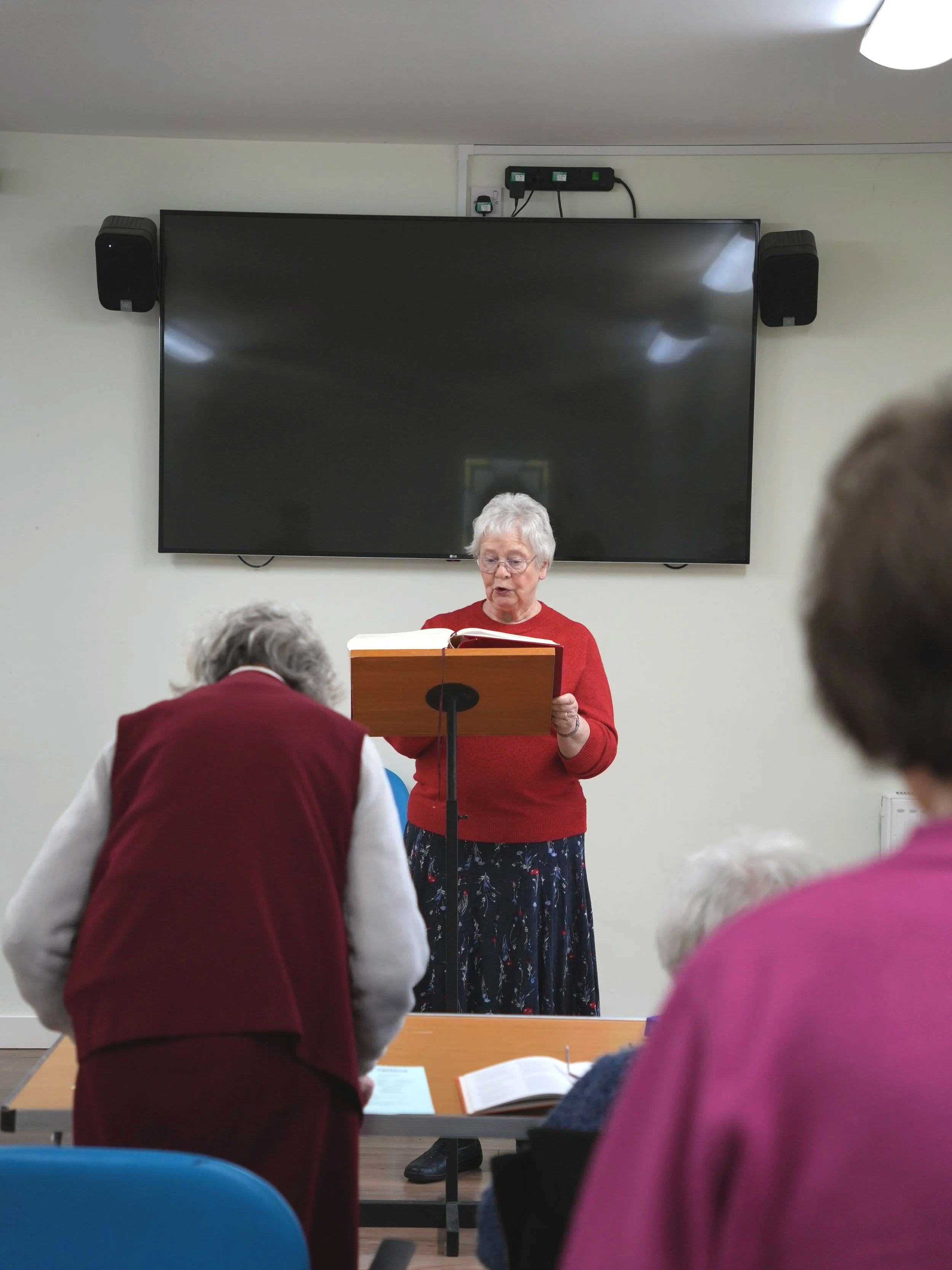An elderly woman standing at a lectern reading from a book, with elderly people seated in front of her in a room with a large TV screen behind her.