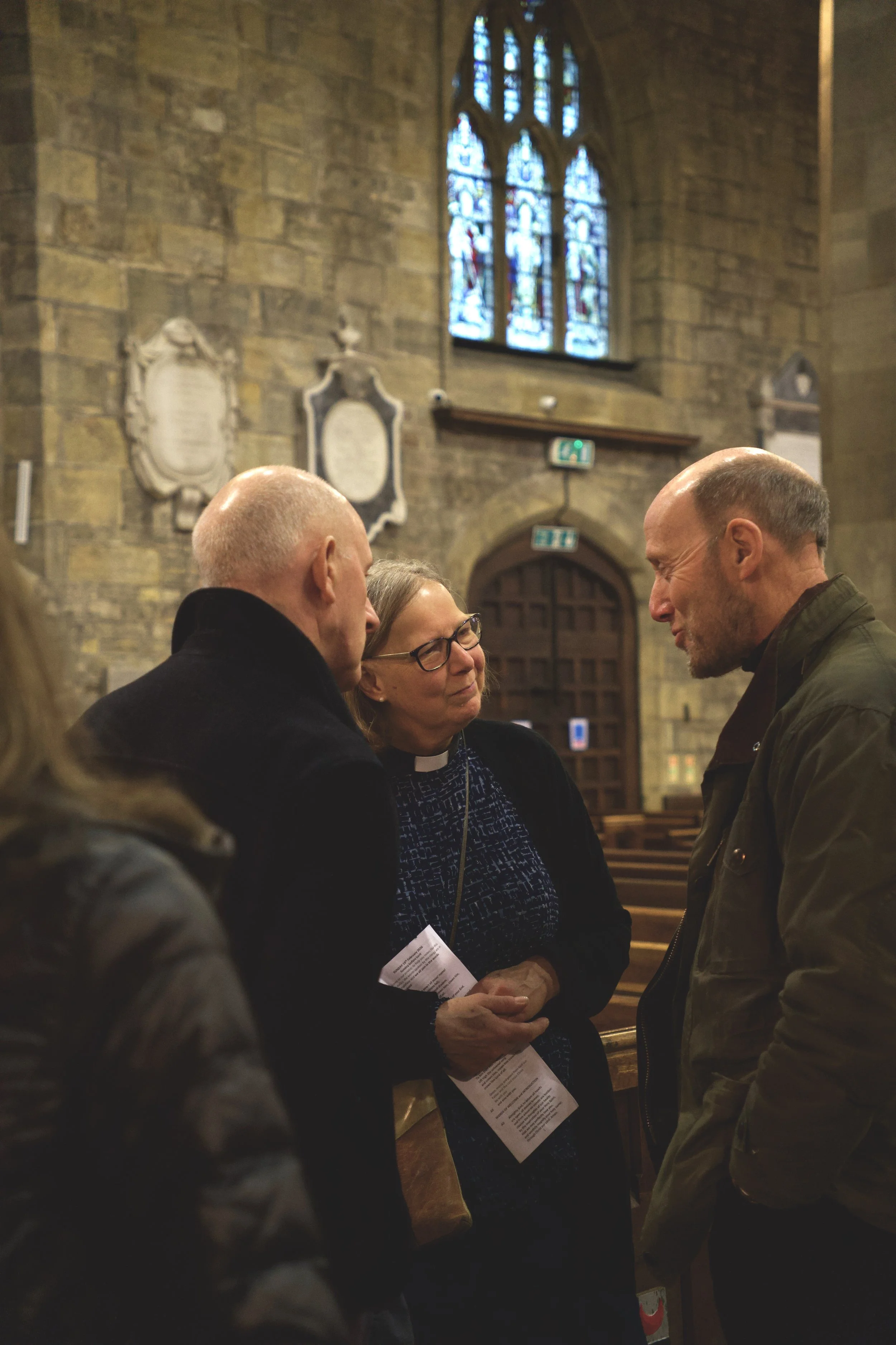 A woman dressed as a clergy member and others are engaged in a discussion inside a church with stained glass windows.
