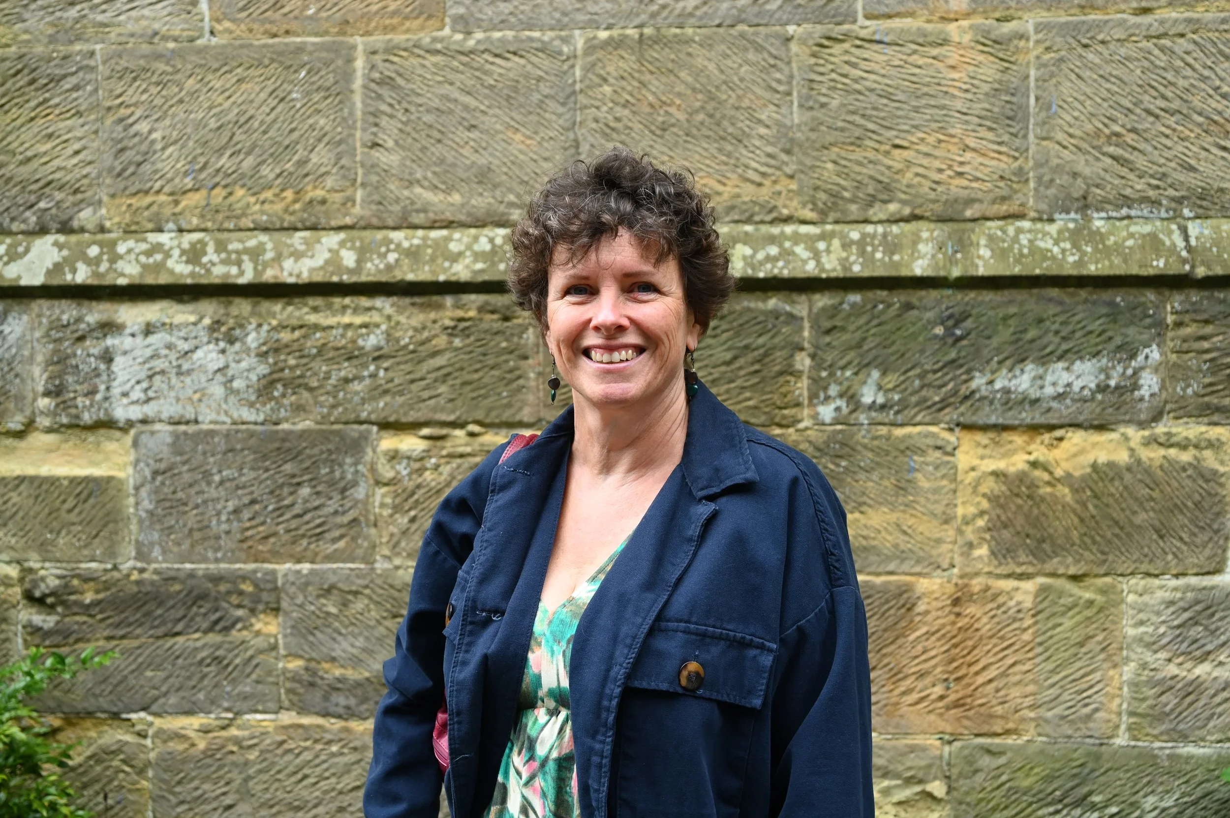 A woman with short curly brown hair is standing outdoors in front of a textured stone wall. She is smiling and wearing a dark blue jacket over a patterned blouse.
