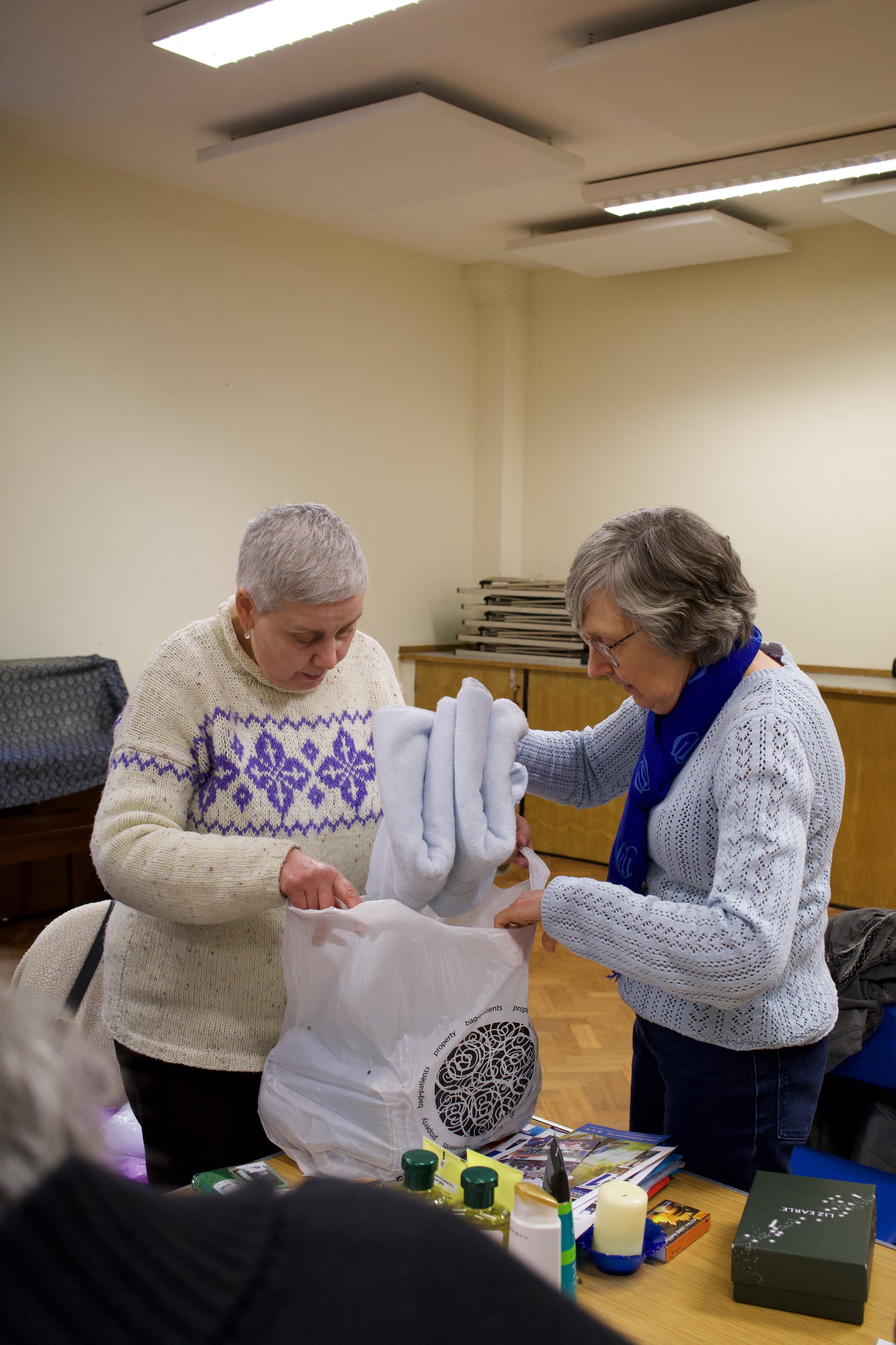 Two women are in a room with a table, looking into a plastic bag that contains a folded white towel or blanket. One woman has short gray hair and is wearing a cream-colored sweater with purple patterns, while the other has gray hair and glasses, dressed in a light gray knitted sweater with a blue scarf. The table has various small bottles, a book, and a box on it.