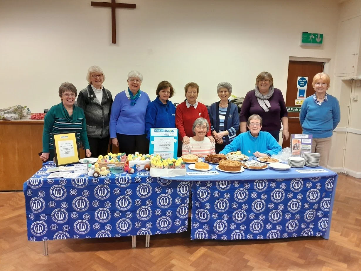Group of women standing behind a table with baked goods and cakes, inside a room with a cross on the wall.