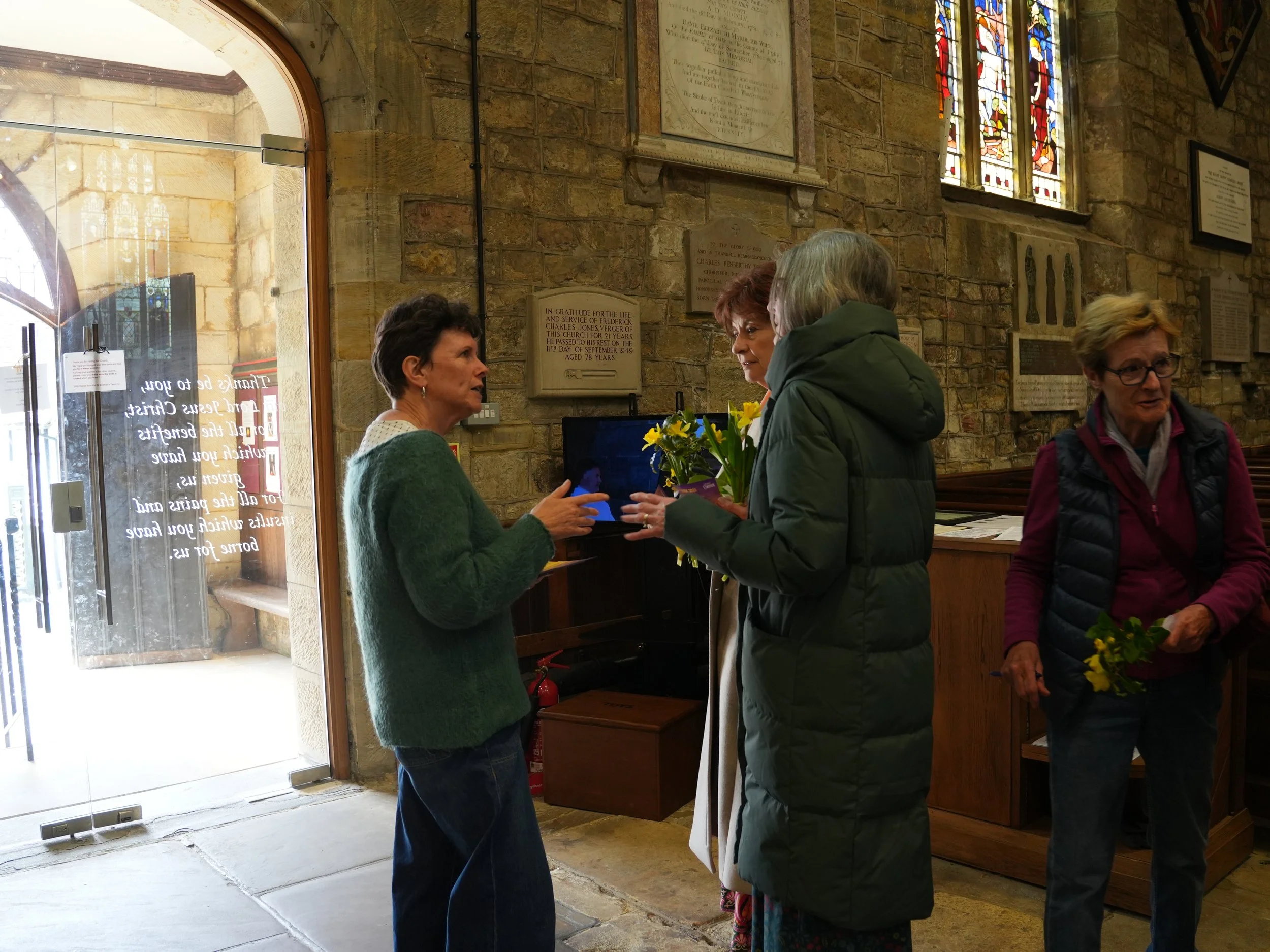 Four women inside a stone church, engaged in conversation. One woman is holding a bouquet of yellow flowers. The church features stained glass windows and plaques on the wall.