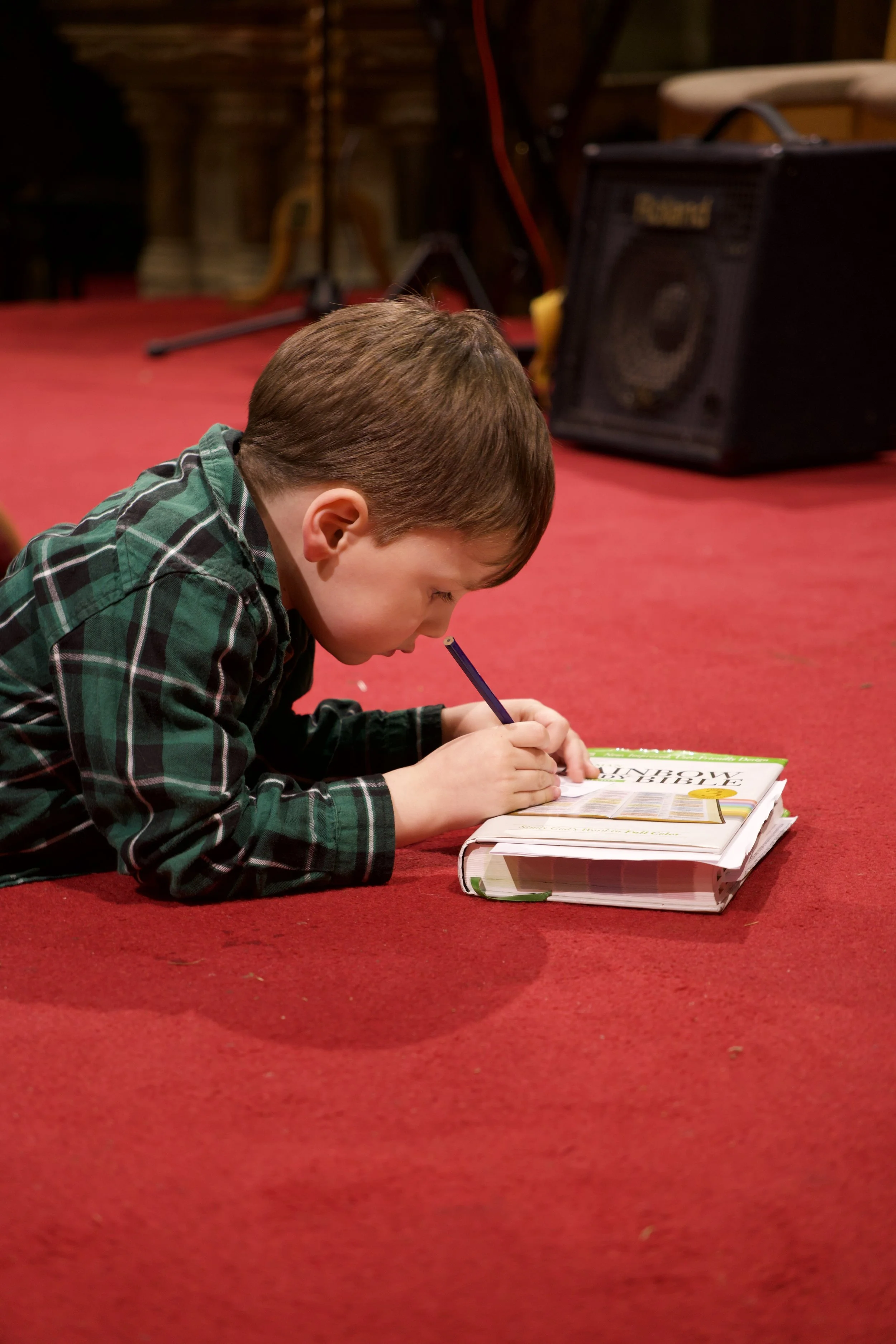 A young boy in a green plaid shirt is lying on a red carpet, writing or drawing in a book with a pencil.