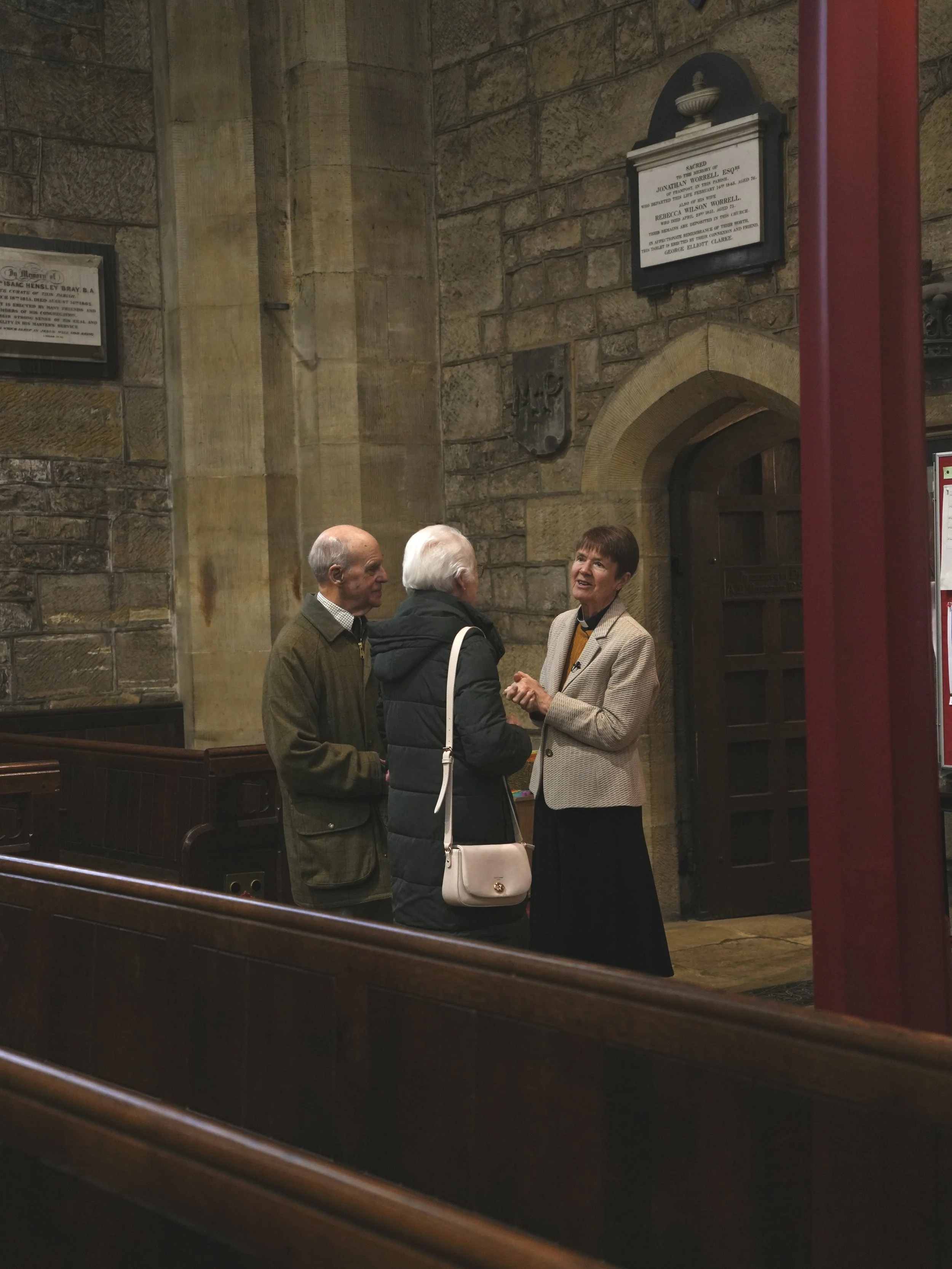 Three people talking inside a church. The church has stone walls and wooden pews. There is a woman with short hair and a beige jacket, and two men, one with gray hair and one with white hair, wearing dark coats. The woman appears to be explaining something to the two men.