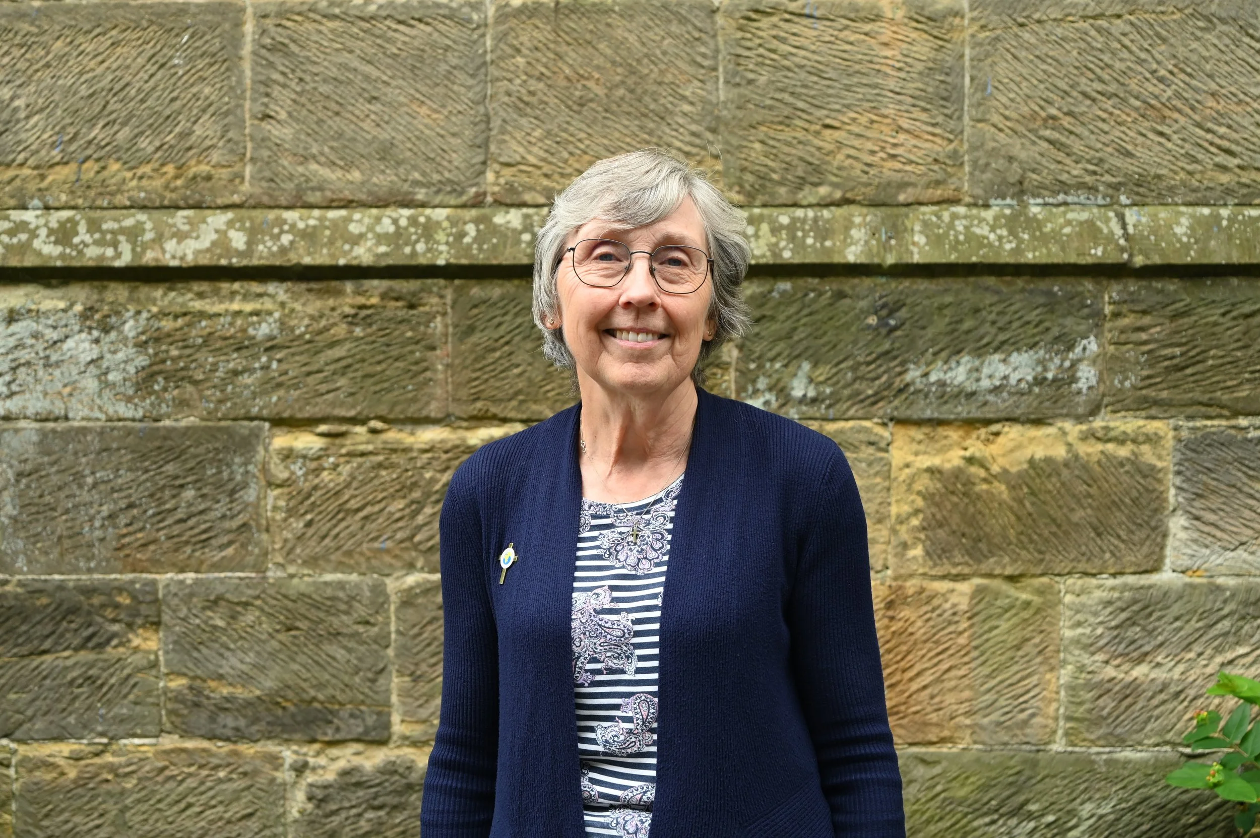 An elderly woman with short gray hair, glasses, and a friendly smile, standing outdoors in front of a stone wall, wearing a dark blue cardigan over a patterned blouse.