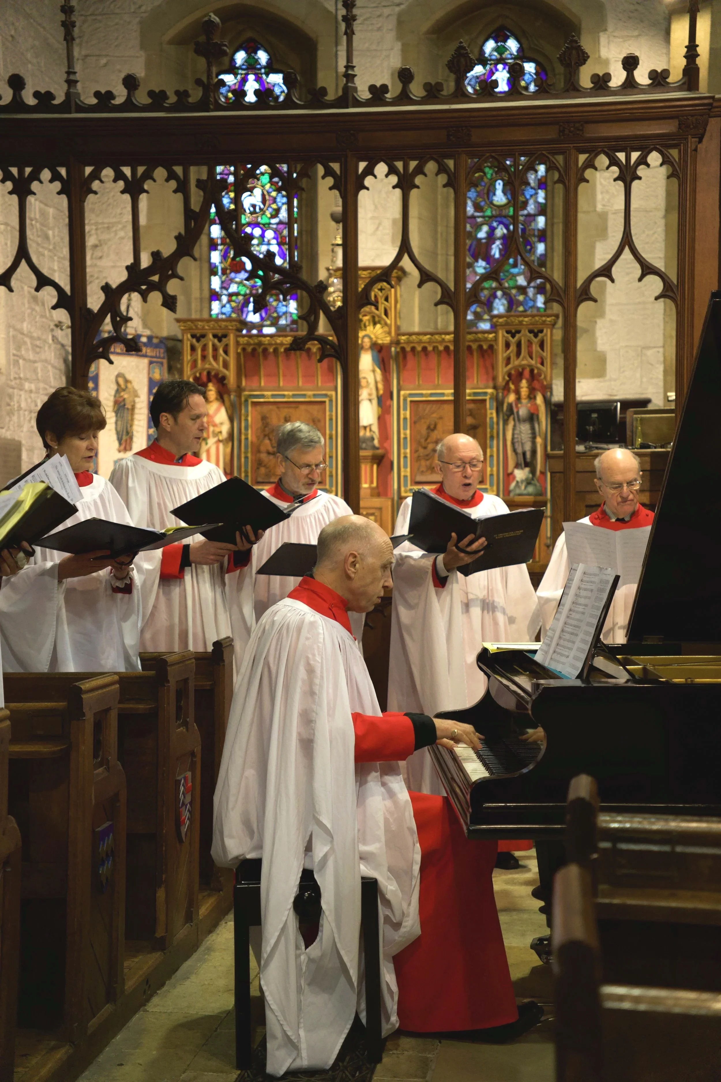 A choir of seven people singing in a church with a pianist playing the piano. The church interior has stained glass windows and religious statues in the background. The choir members are dressed in white robes with red accents.