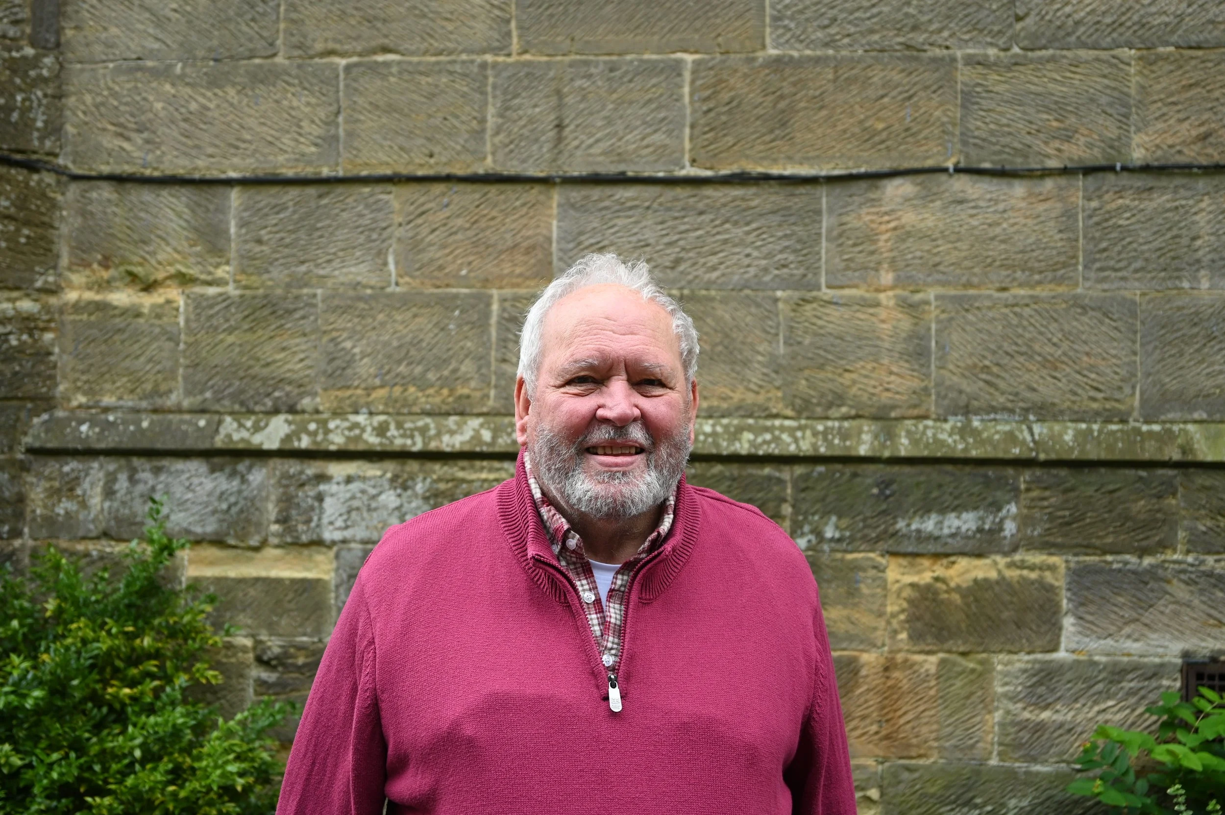Older man with white hair and a beard wearing a maroon sweater and checkered shirt, smiling outdoors in front of a stone wall with green bushes.