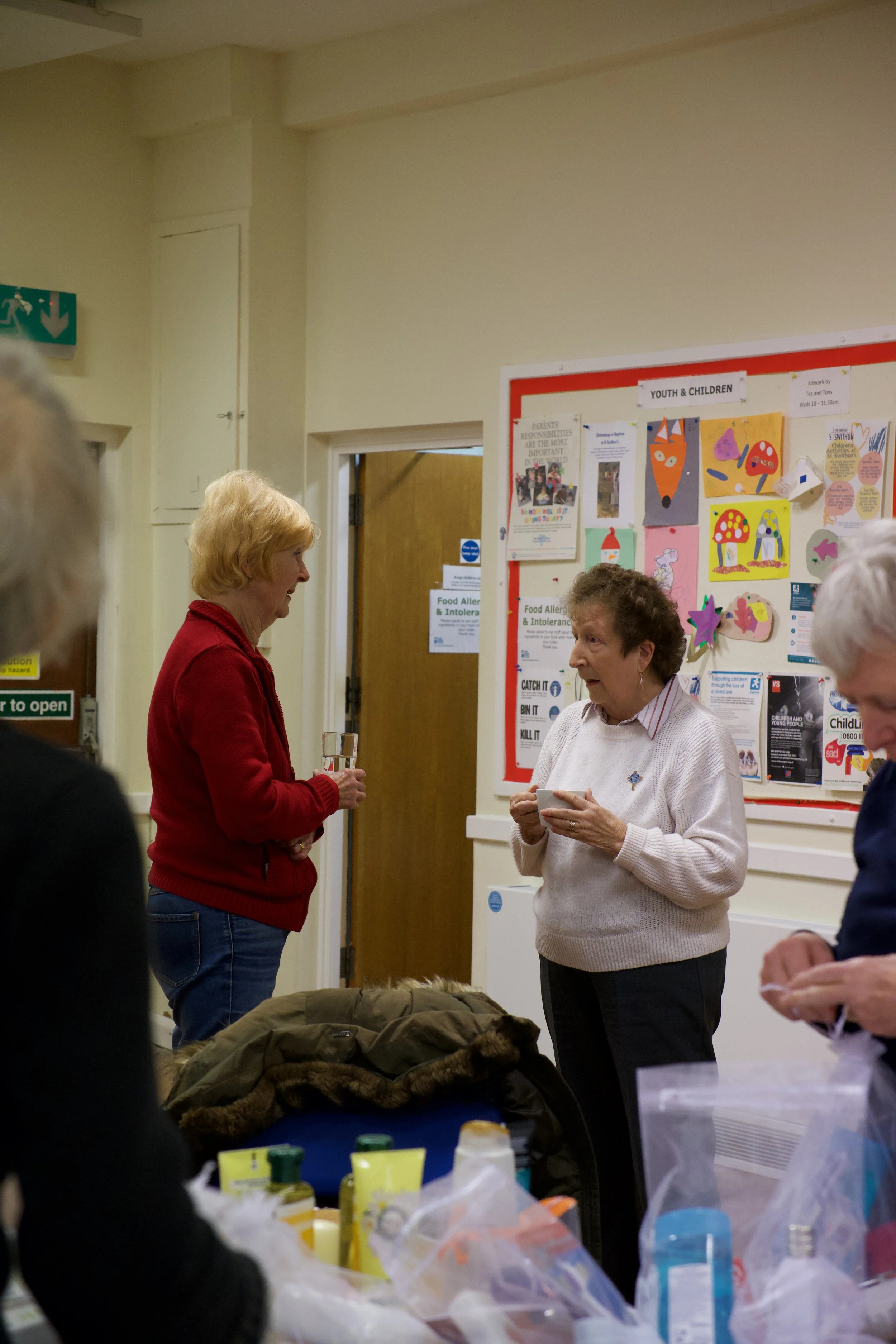 Three women engaging in conversation inside a community center or meeting room, with a bulletin board titled 'YOUTH & CHILDREN' in the background and various papers and artwork pinned on it. One woman is holding a cup, and another is holding a glass of water. The scene includes a table cluttered with various supplies and a coat placed on a chair.