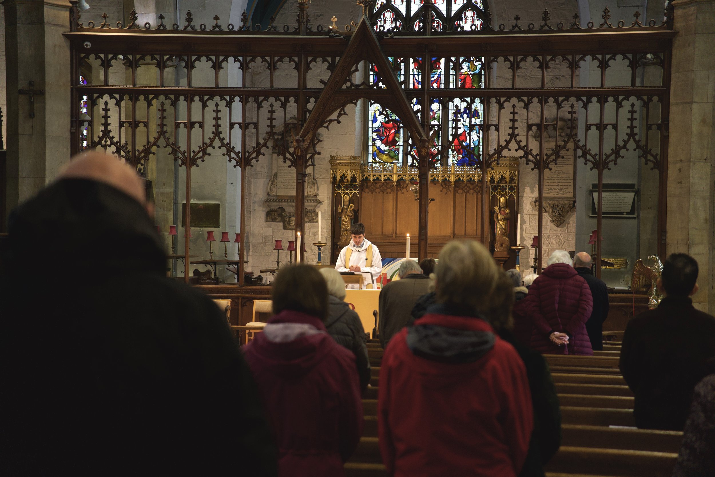 People gathered in prayer or reflection inside a church, listening to a priest or clergy member reading at the altar, with stained glass windows and ornate woodwork in the background.