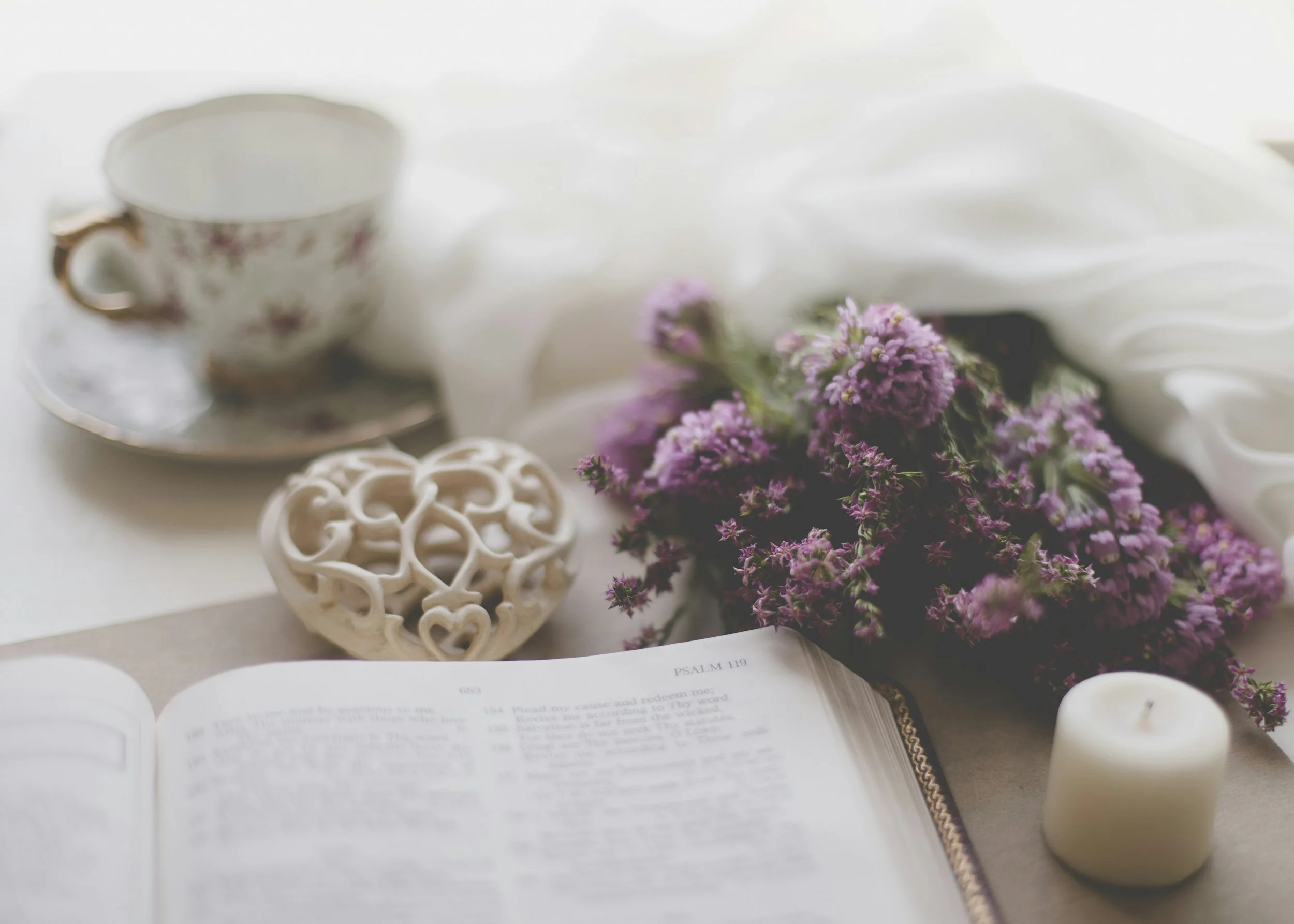 A still life scene with a cup and saucer, a decorative heart-shaped object, a bouquet of purple flowers, an open book, and a white candle on a white surface with soft natural lighting.
