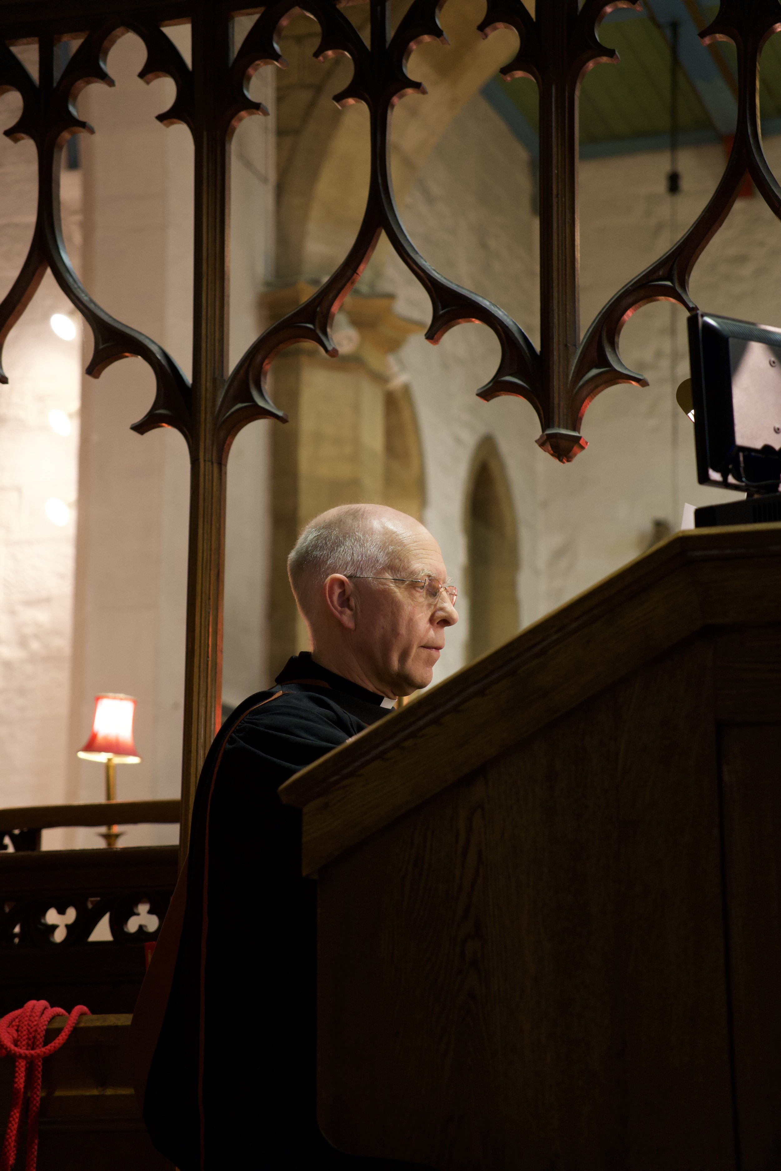 A priest or monk in a dark robe sitting at a wooden lectern inside a church with stone walls and arched windows, behind decorative ironwork.