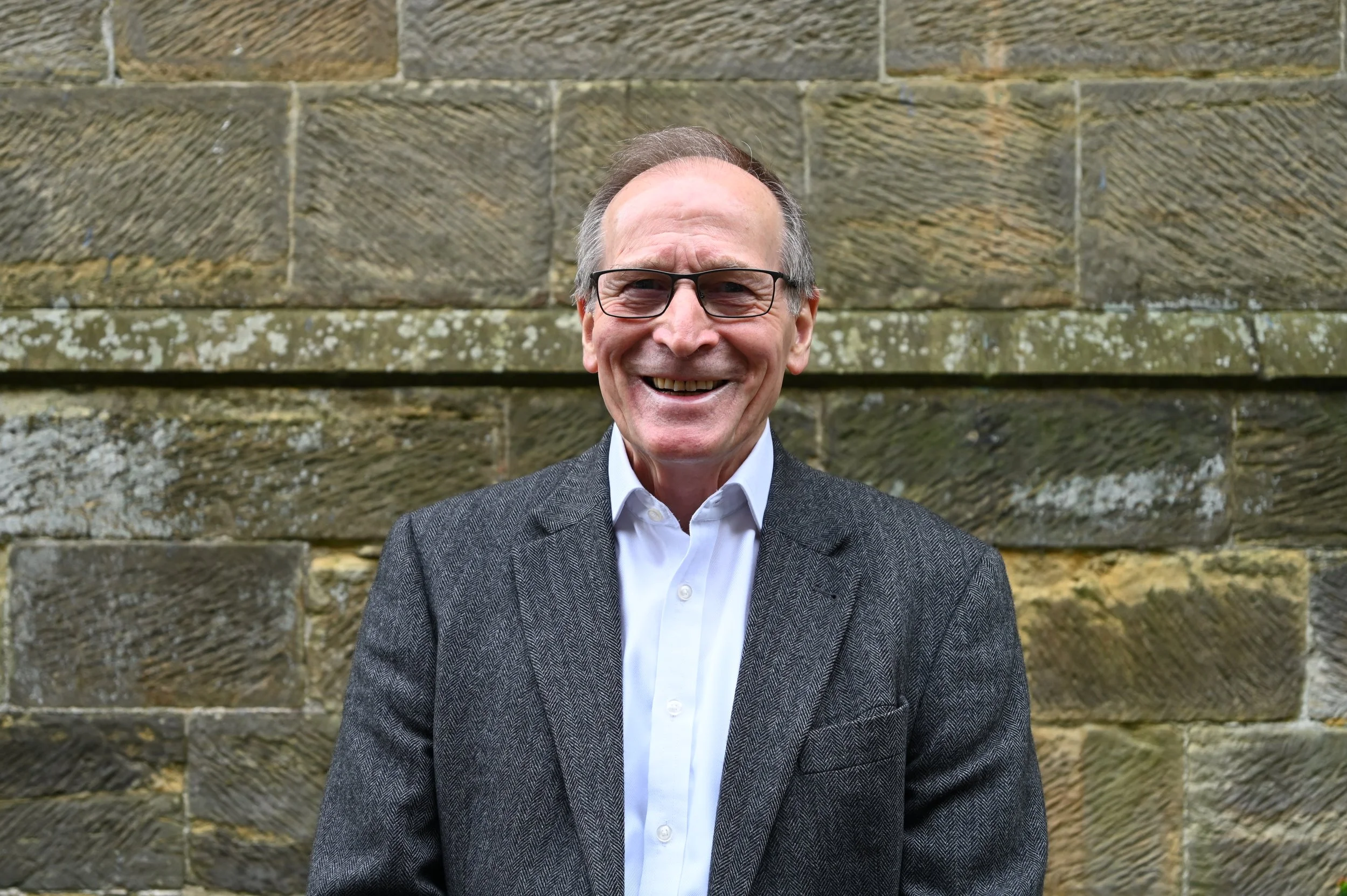 A smiling man wearing glasses, a white dress shirt, and a gray suit jacket standing in front of a stone wall.