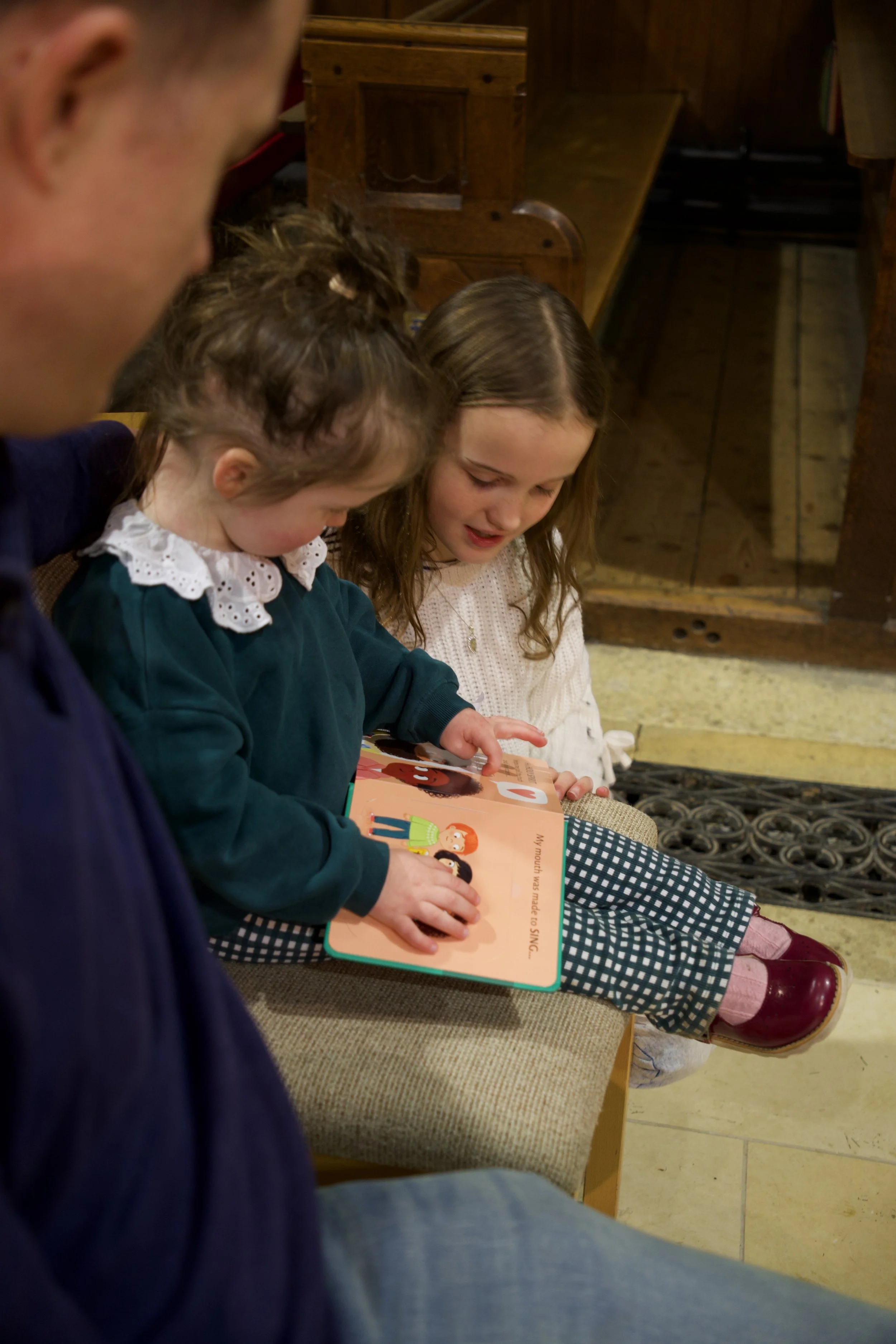 Two young girls sitting on a bench, reading a children's book, with an adult partially visible in the foreground. The girls are focused on the book, which has colorful illustrations.