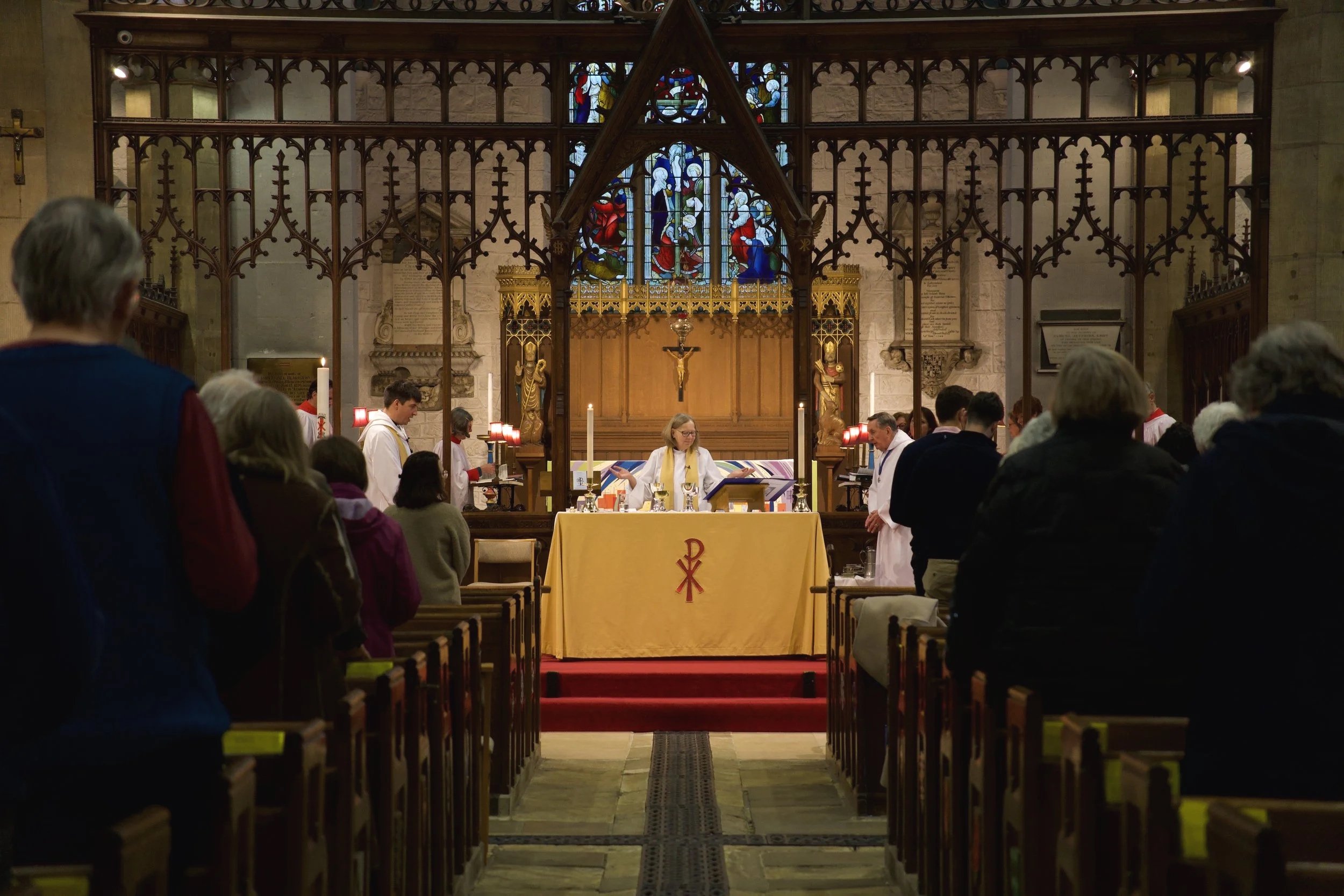 A church interior during a religious service with a priest at the altar, surrounded by congregation members, some dressed in liturgical vestments, near stained glass windows.