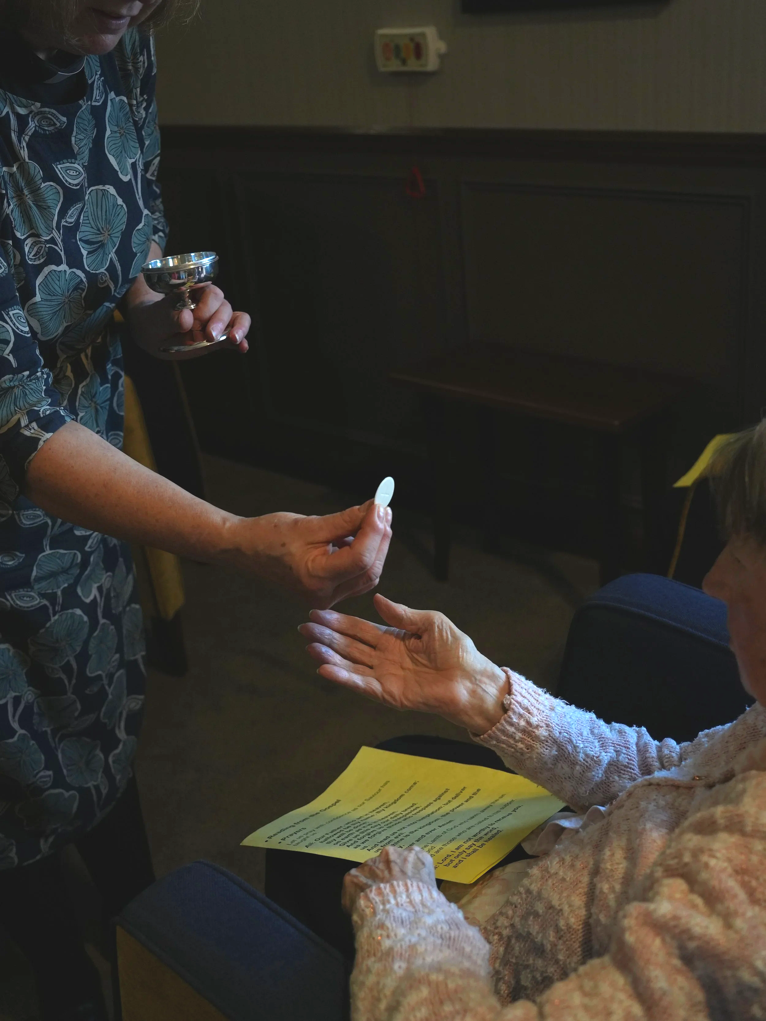 A woman hands a pill to an elderly woman sitting in a chair, with a paper on her lap and a small dish in her other hand. The setting appears to be a medical or care facility.