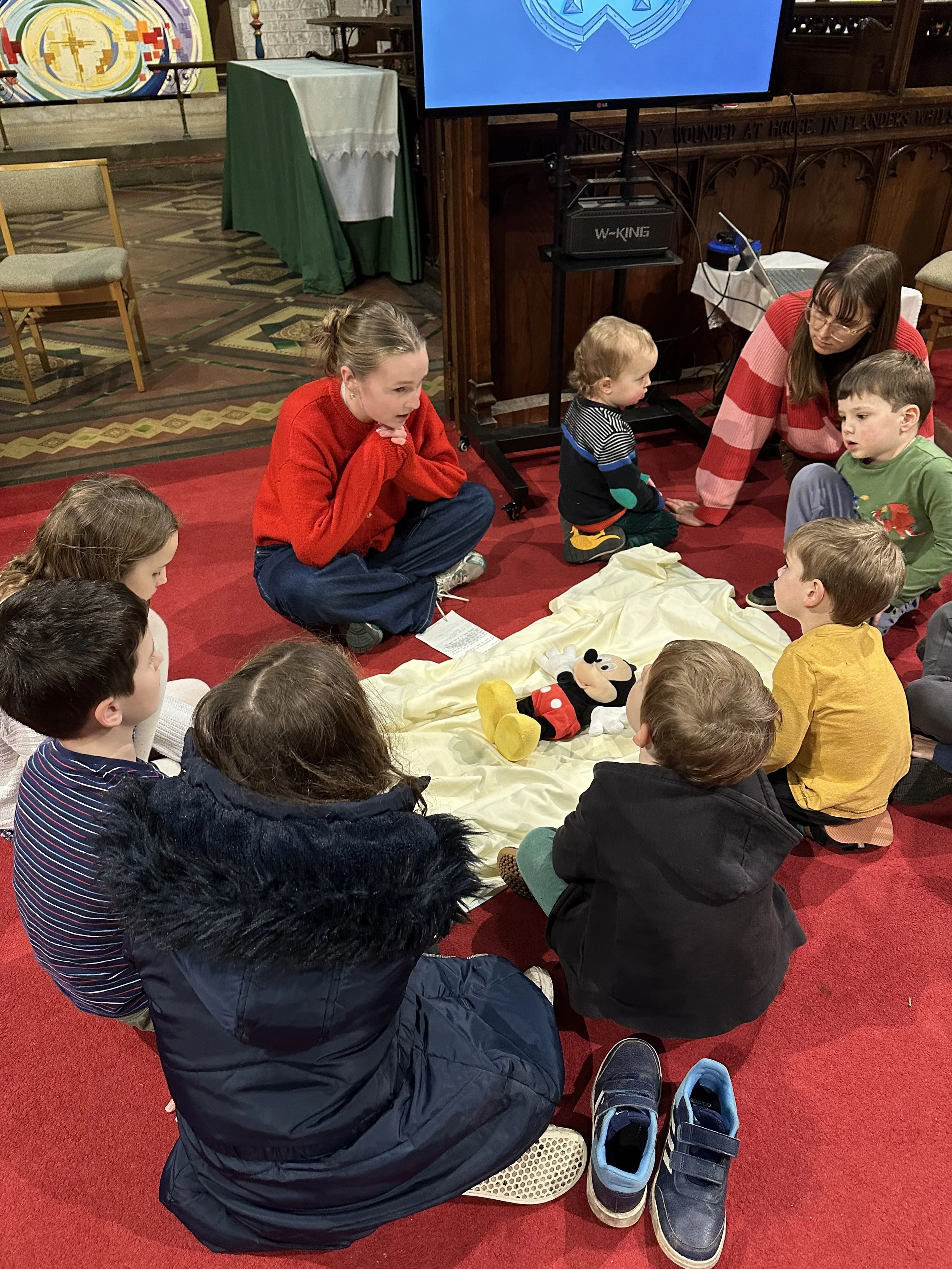 Children and two adults gathered on a red carpeted floor around a Mickey Mouse plush toy on a cream-colored blanket, watching a presentation or activity in a church or community hall.