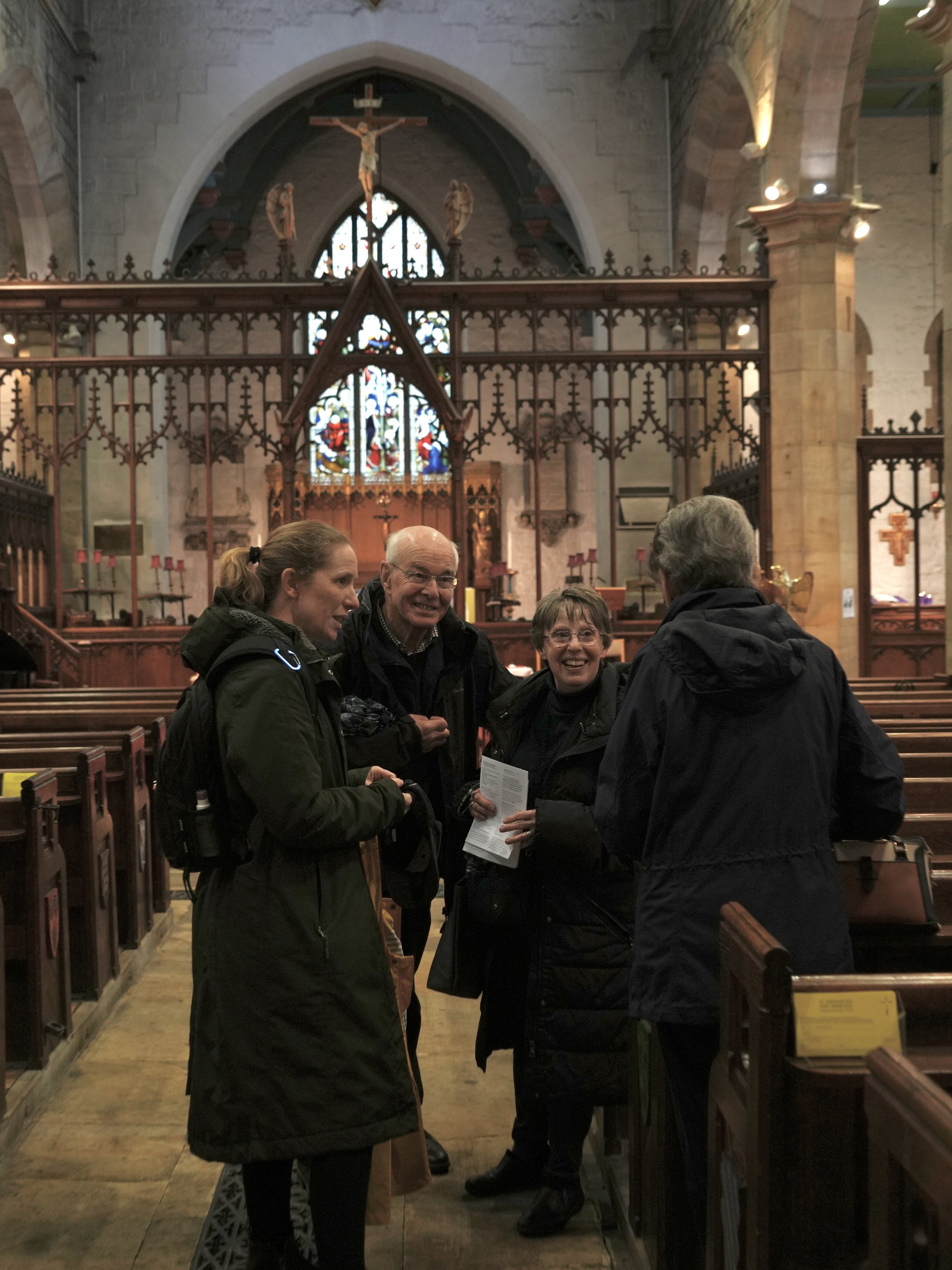 Four people standing and chatting and smiling inside a church with wooden pews, stained glass windows, and religious statues and symbols in the background.