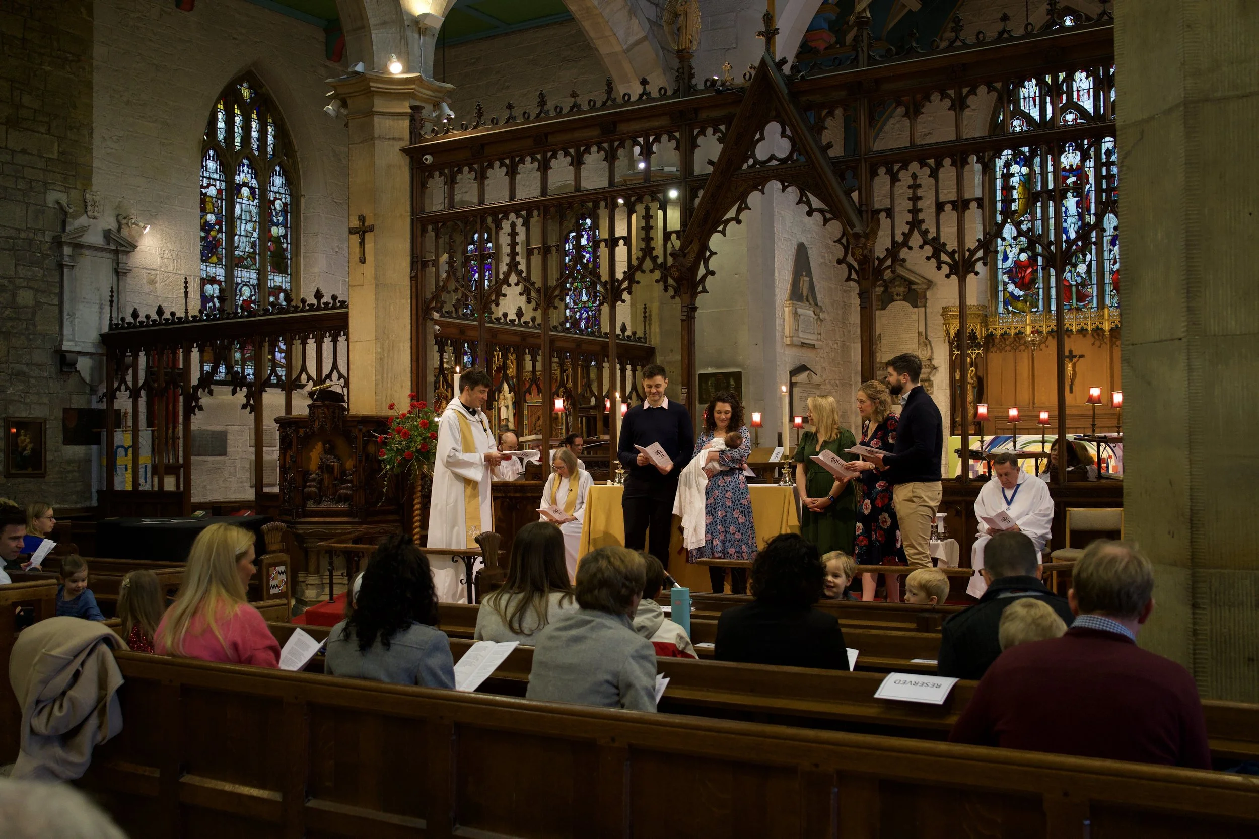 A baptism ceremony taking place inside a church, with a priest, family members, and congregation, in front of stained glass windows and wooden church architecture.