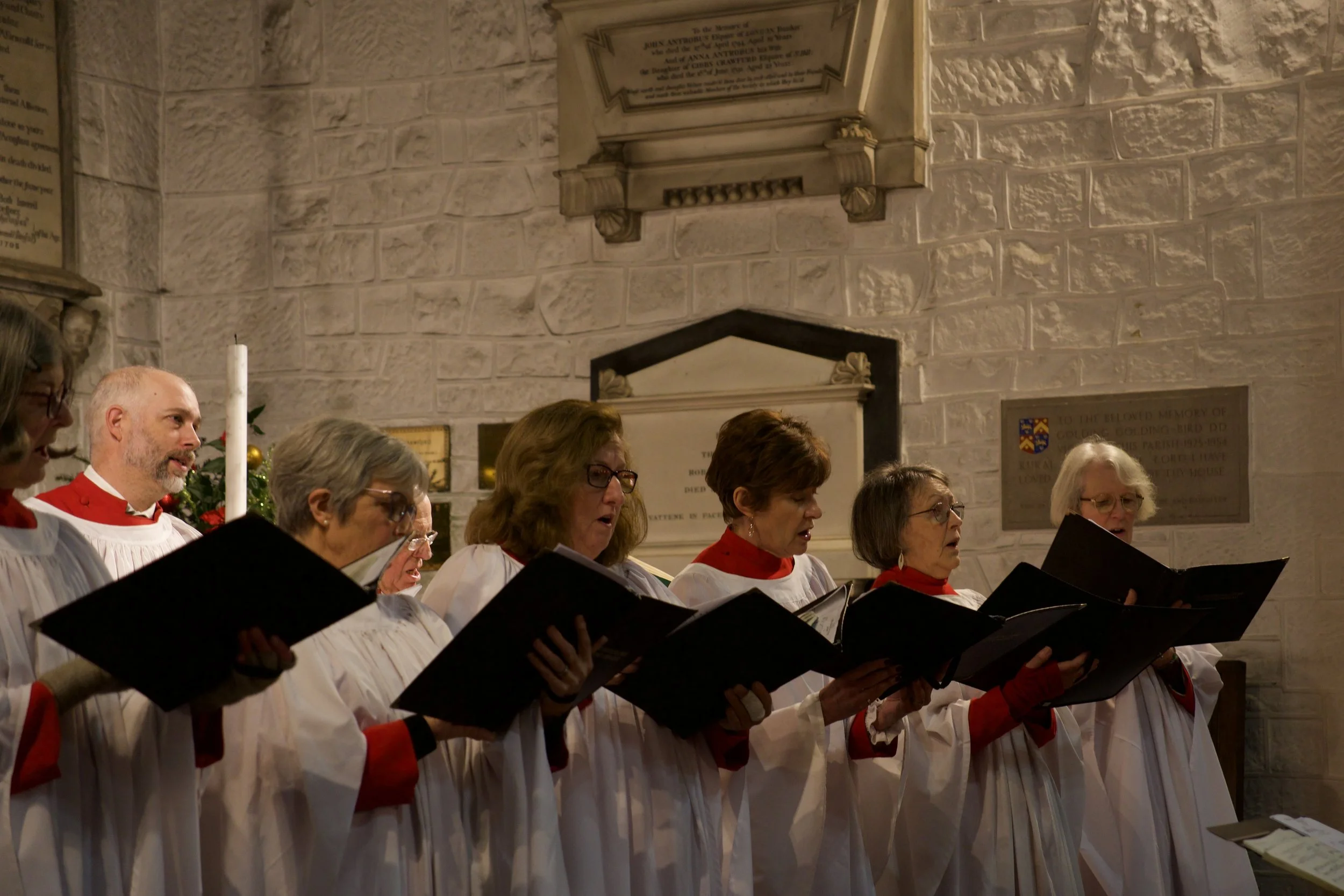 A choir of men and women singing in a church, holding black songbooks, dressed in white robes with red accents, in front of stone walls and memorial plaques.