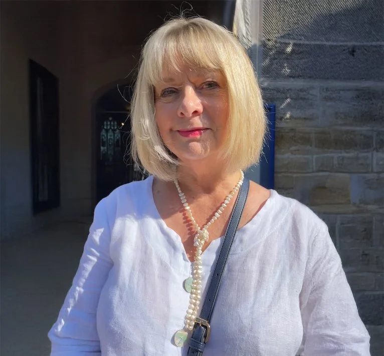 A woman with blonde hair wearing a white top, pearl necklace, and purse, standing outdoors in sunlight.