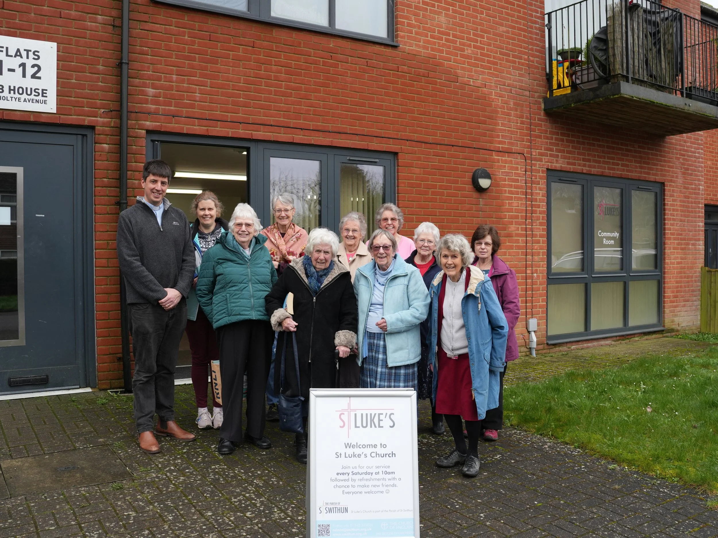 Group of elderly women and two younger adults standing outside St Luke's Church, smiling. There is a signboard in front welcoming visitors for Sunday service. The church building is made of red brick with black-framed windows and doors. The setting appears to be in a residential area with a grassy and paved area.