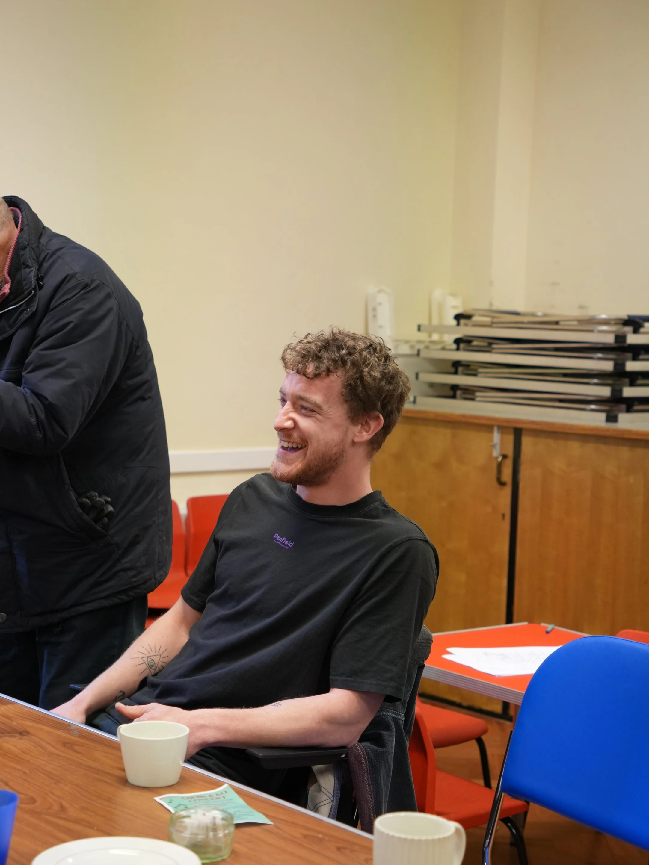 A young man with curly hair and a beard sitting at a table, smiling and laughing, wearing a black t-shirt. There are cups, papers, and a blue chair on the table.