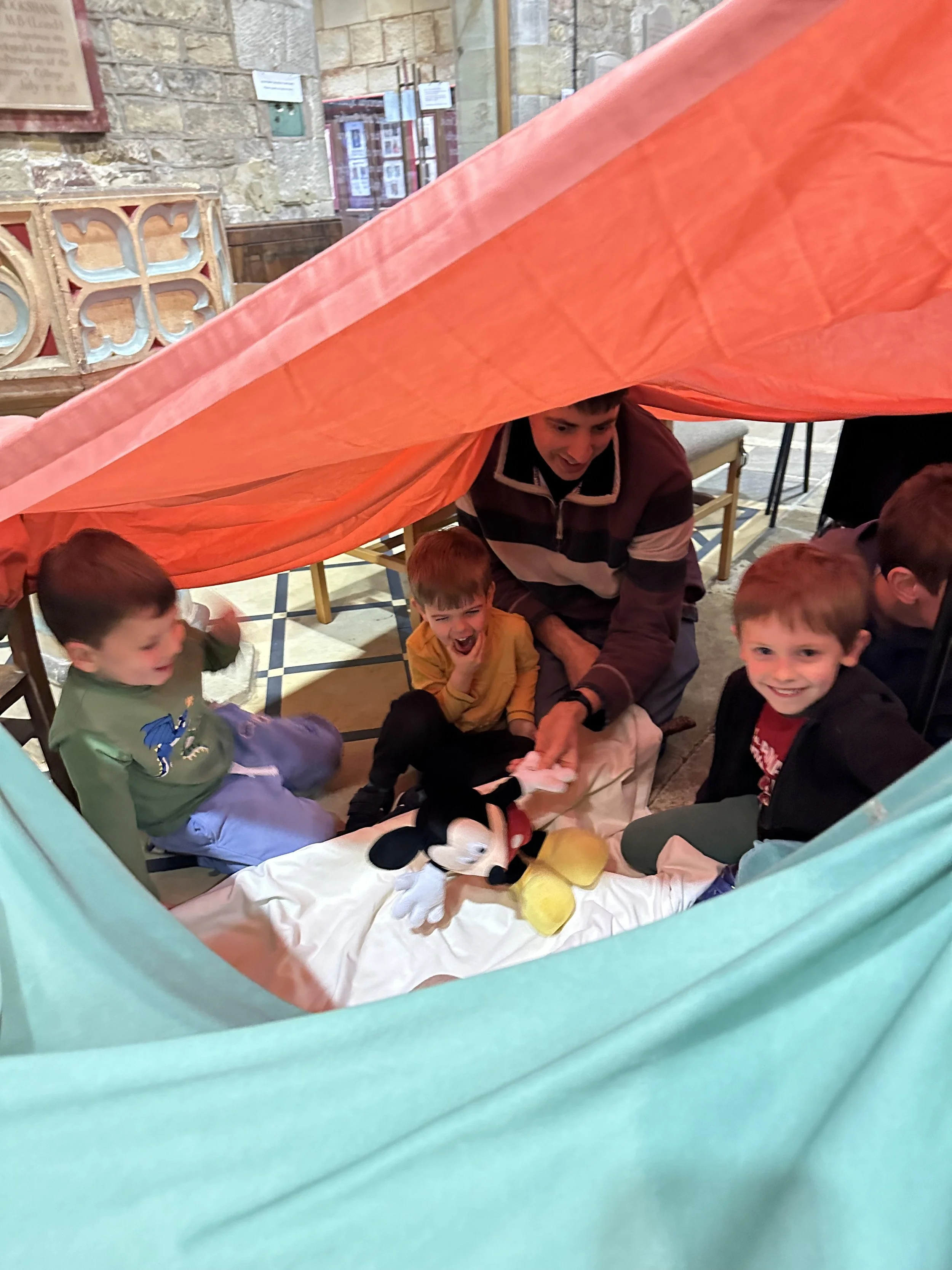 Children and an adult sitting under a colorful cloth tent, playing with a Mickey Mouse plush toy at an indoor location with stone walls and wooden benches.