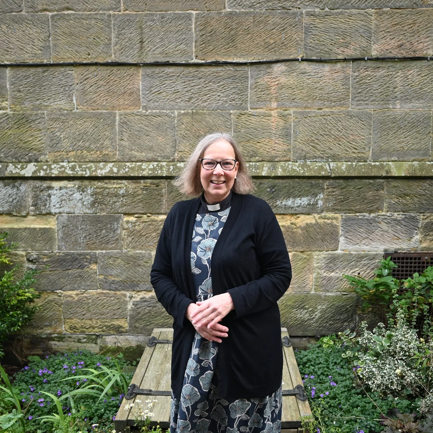 A woman with glasses and shoulder-length gray hair standing outdoors in front of a stone wall, smiling, wearing a black coat over a floral dress, with greenery and flowers around her.