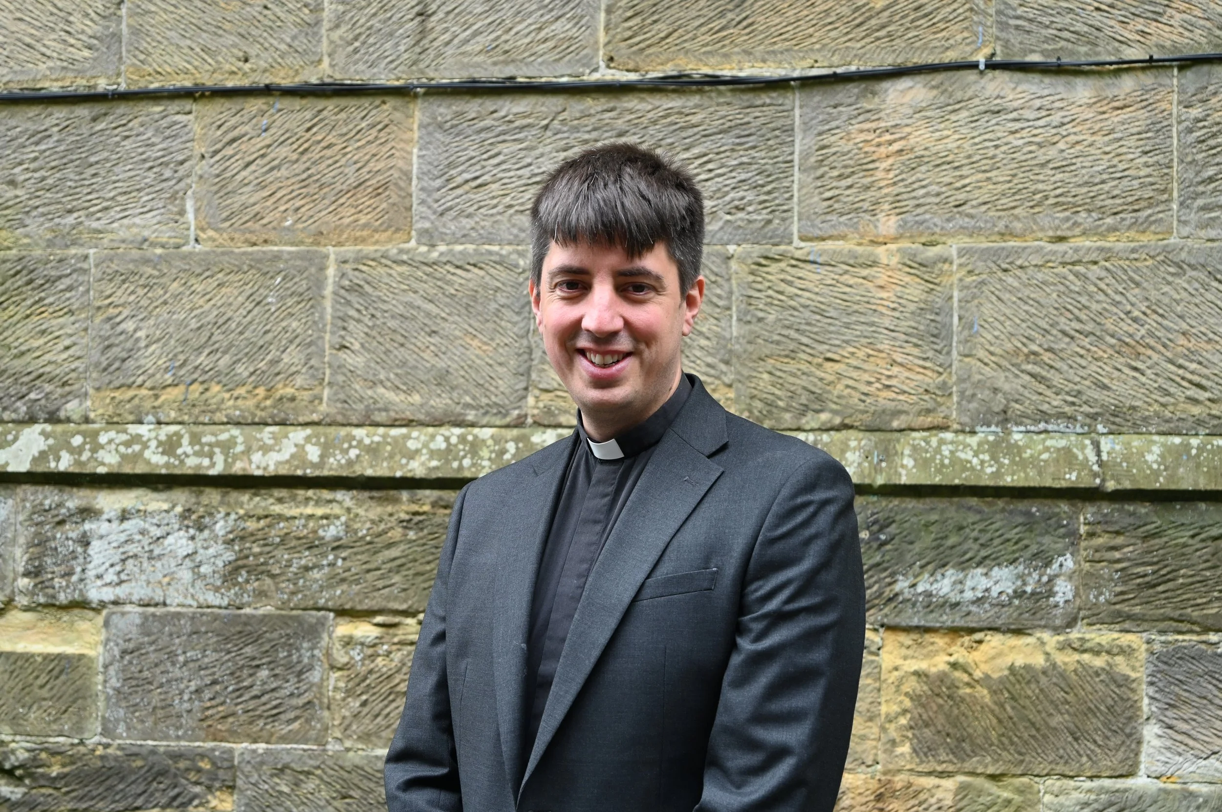 A man in a clerical collar and black suit standing outdoors against a textured stone wall, smiling at the camera.
